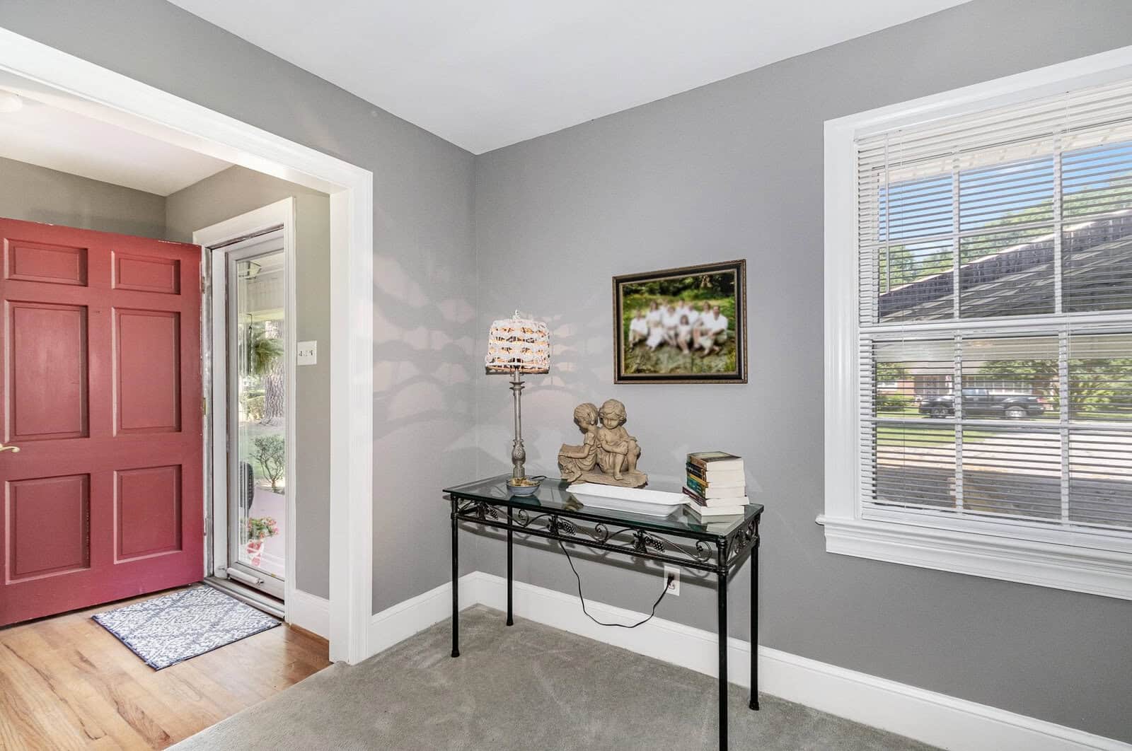 A well-lit entryway featuring a red front door, a small console table with a lamp, books, and a sculpture, against a gray painted wall with a framed picture hanging above it, next to a window with a view of the exterior.