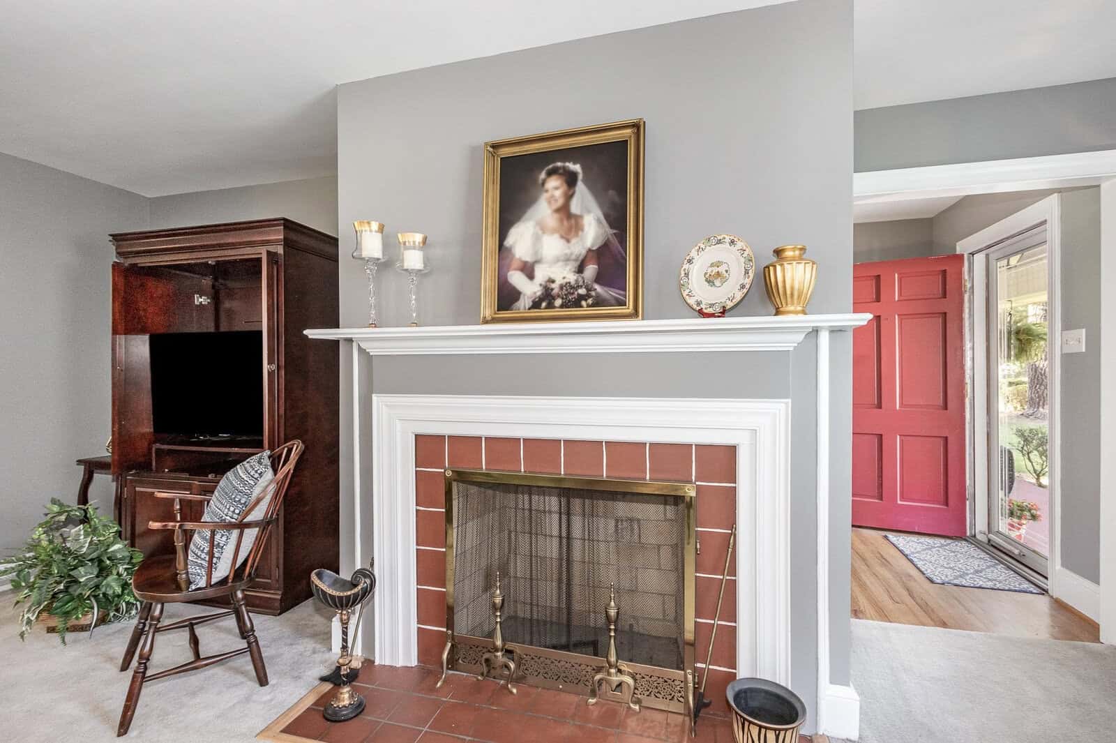 A neatly organized living room featuring a white mantelpiece with a classic portrait above it, flanked by vintage decorative items and a rocking chair to the side; in the background, a vibrant red door offers a pop of color.