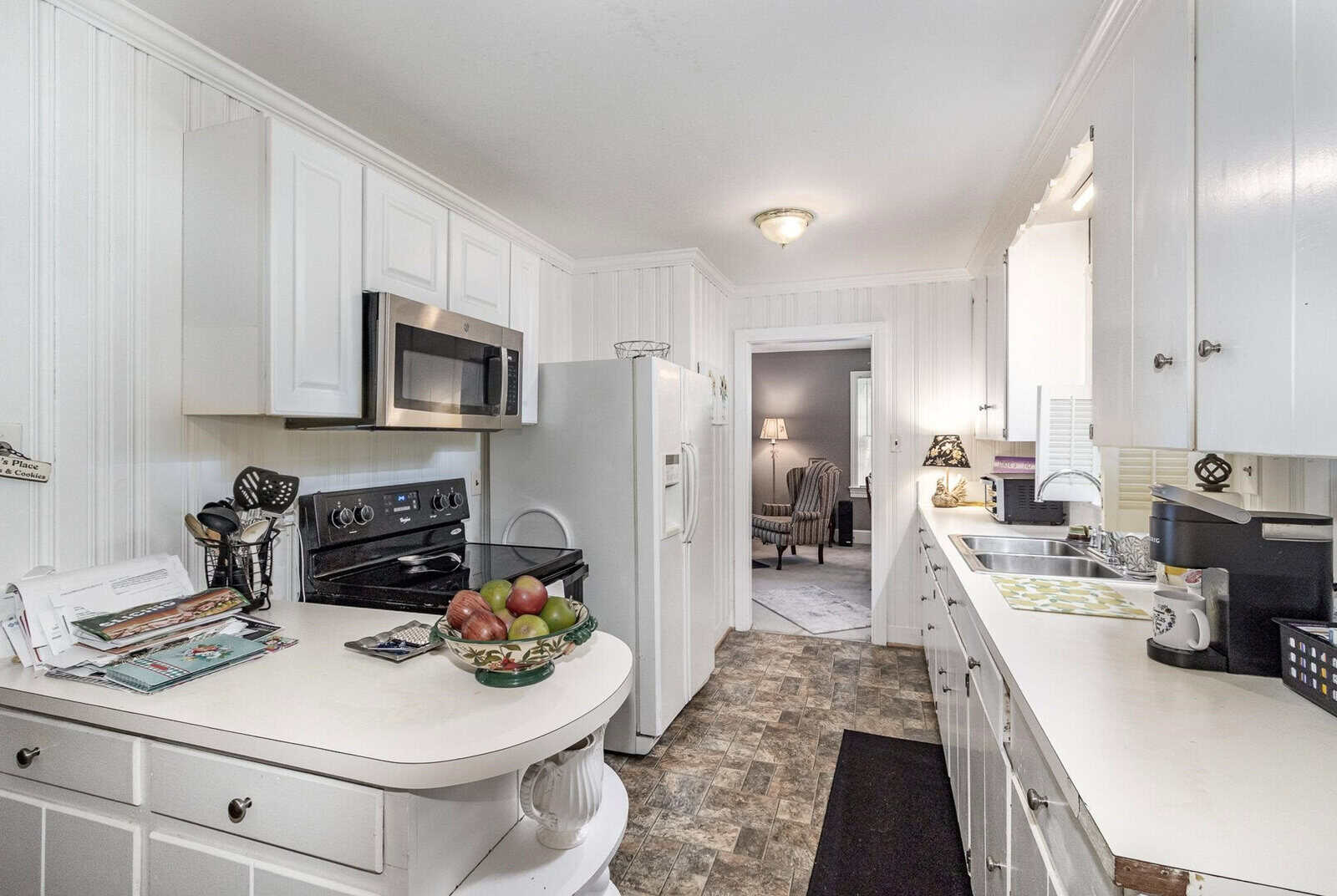 A small, white kitchen with modern appliances, including a microwave and stove, featuring a central island with a bowl of fruit and assorted papers, leading to a hallway and an adjacent room.
