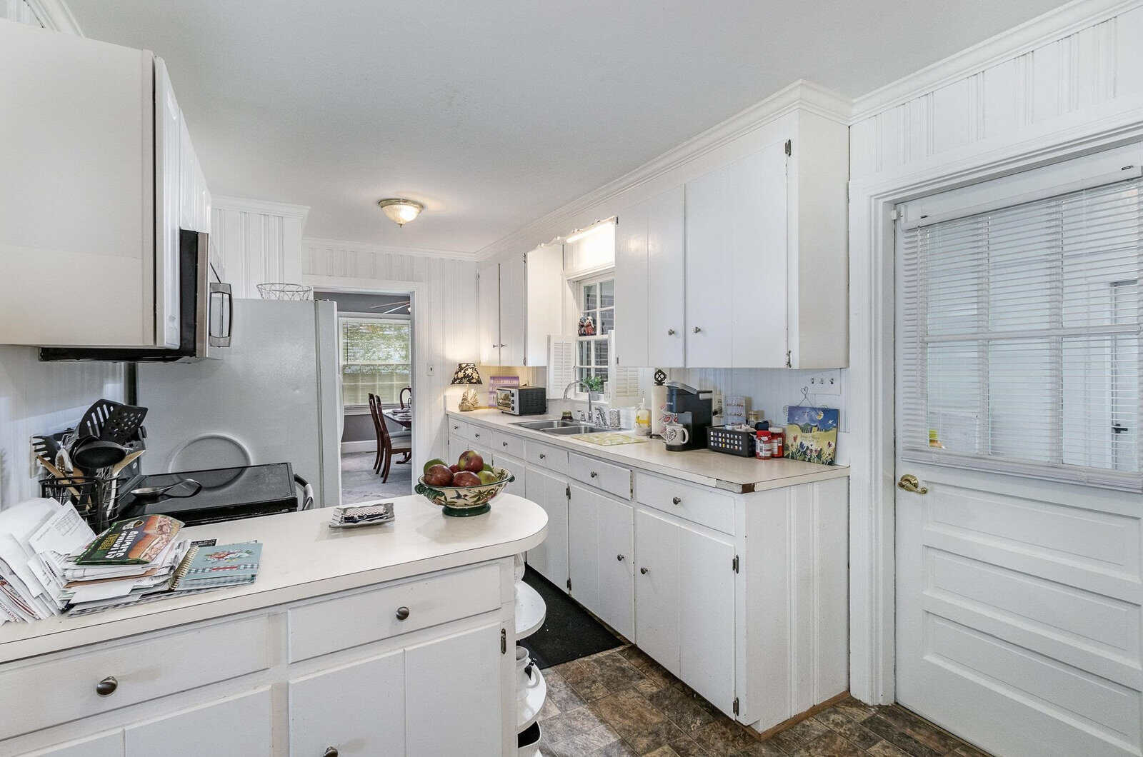 A bright kitchen with white cabinetry and appliances, featuring a window above the sink, a bowl of fruit on the counter, and various kitchen utensils and items arranged throughout the space.