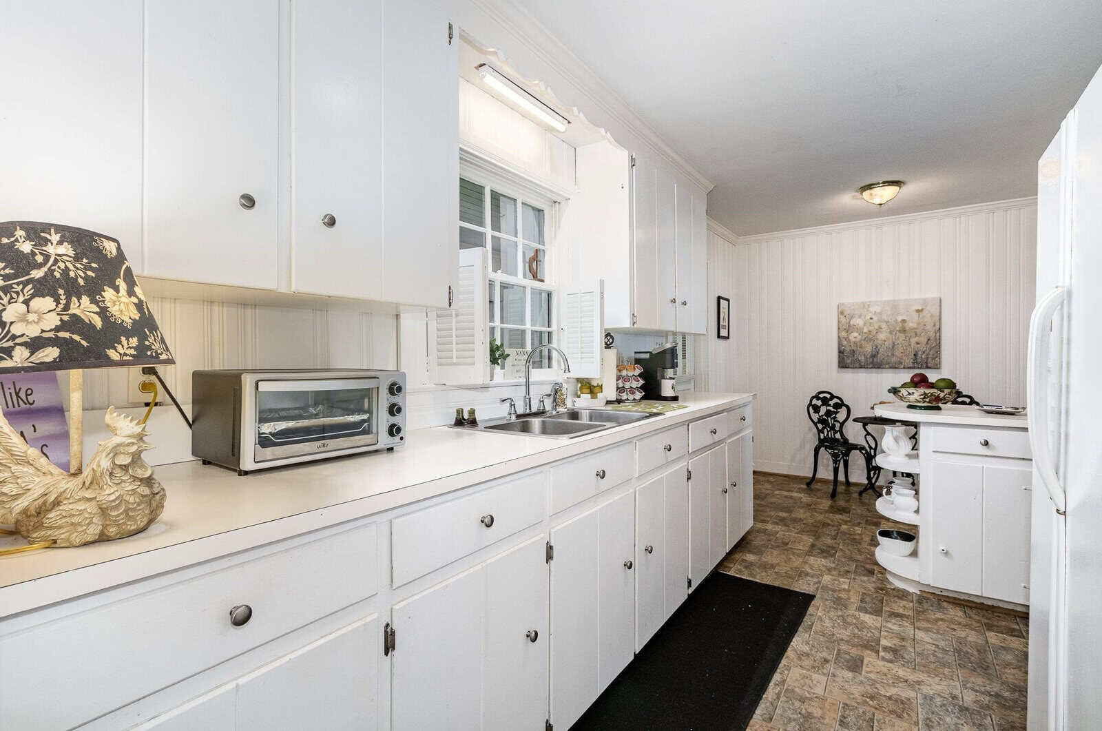 A traditional kitchen interior with white cabinetry, a microwave on the countertop, a window above the sink, and a small dining area with a round table and chairs.