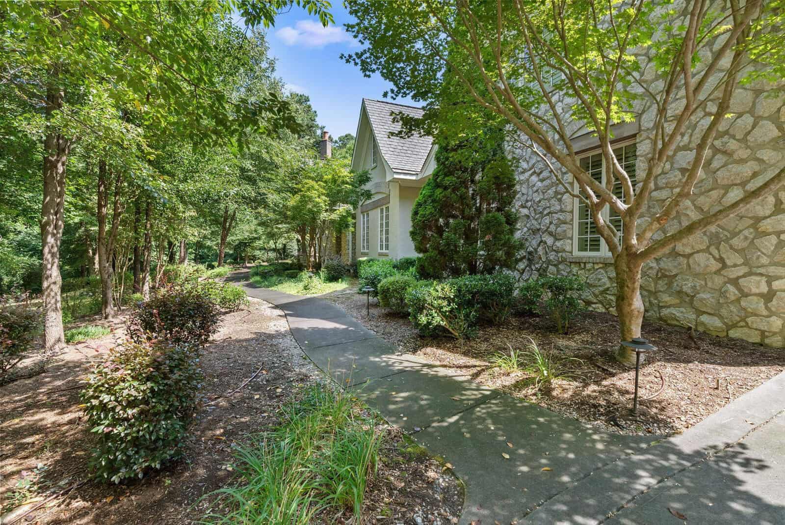 A stone house with a steeply pitched roof is nestled among lush greenery with a winding path leading to the entrance.