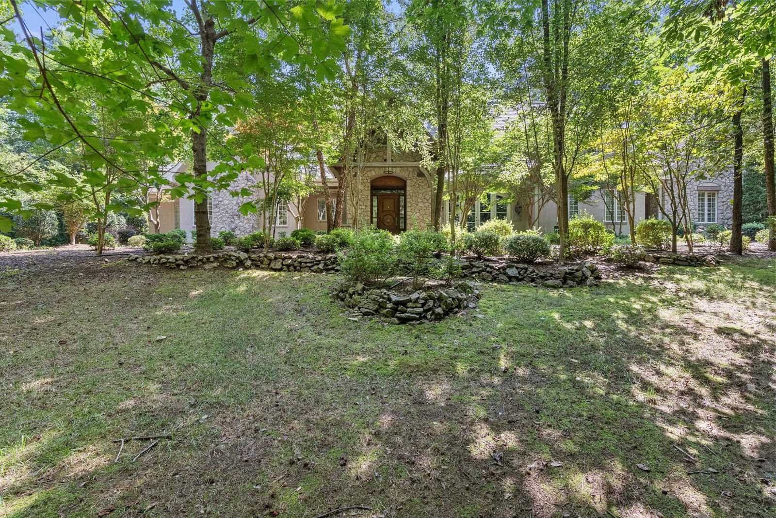 A suburban brick house partially shaded by trees, featuring a central front door and a landscaped garden with a rock border.