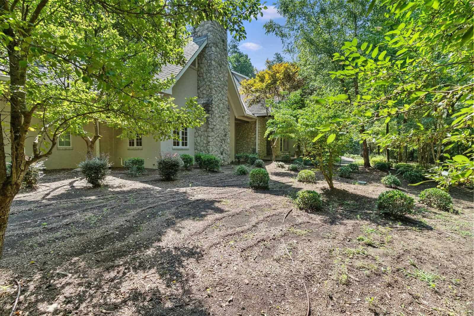 A single-story house with a stone facade is partially obscured by green foliage in a garden setting with freshly tilled soil.