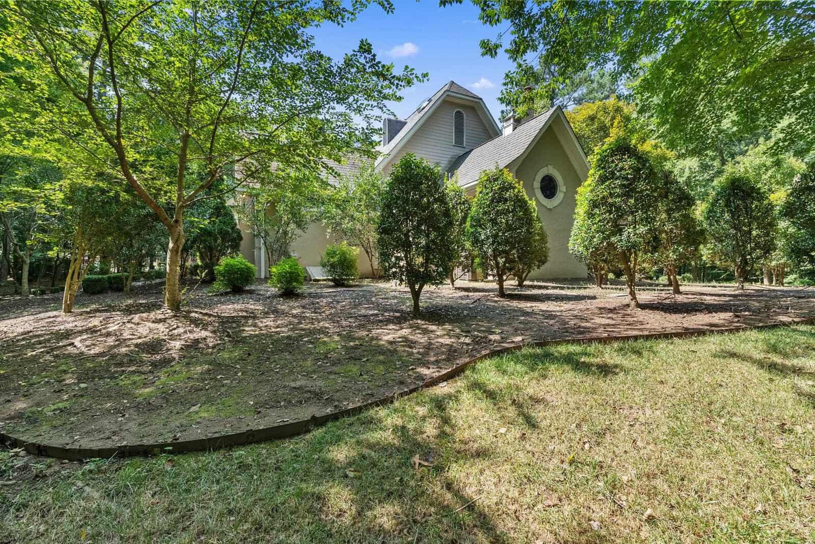 A spacious residential backyard with manicured grass, several trees, a retaining border, and the back side of a modern house with large windows.