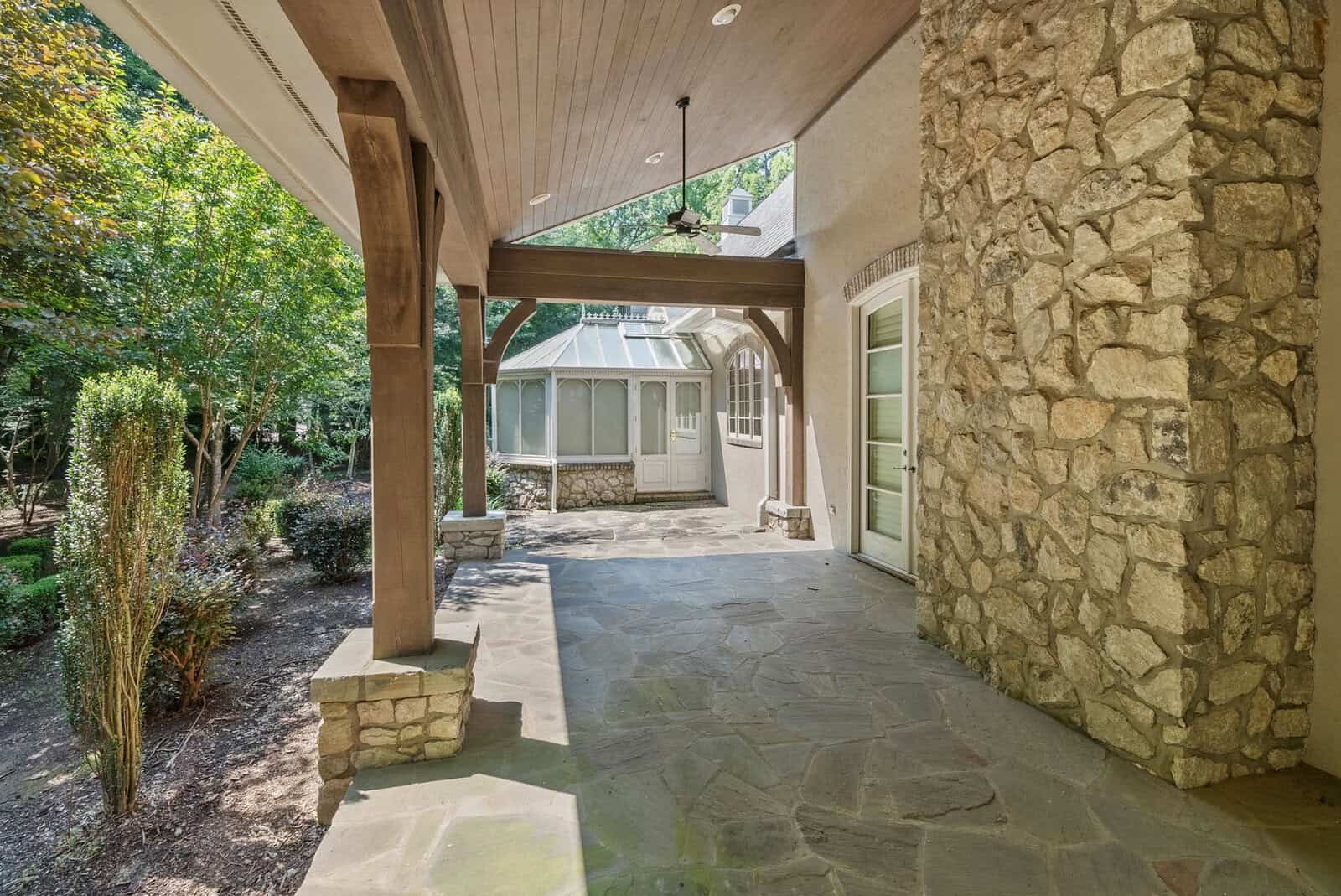 Covered stone patio area leading to a house with white doors and windows, surrounded by greenery.