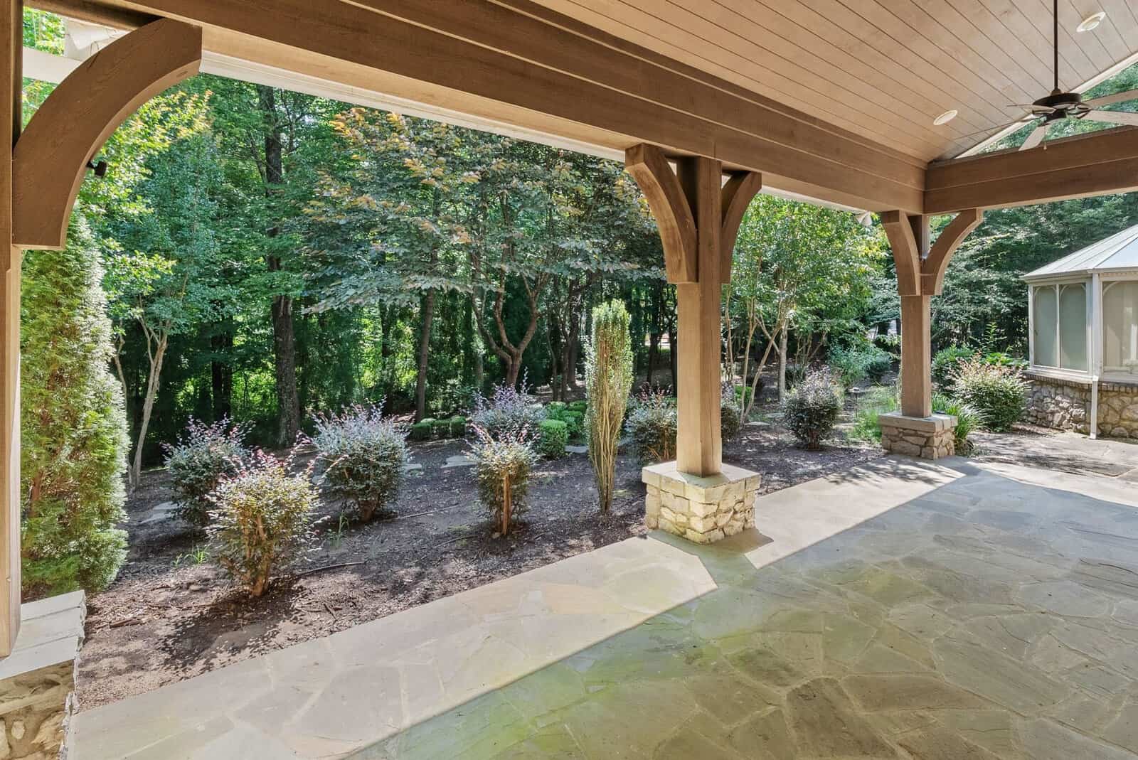 A spacious covered patio with stone flooring overlooks a landscaped backyard with multiple shrubs and trees, offering a view of a natural, wooded area.