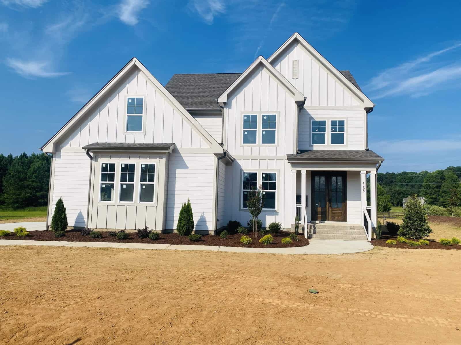 A modern two-story white house at 1200 Pond Side Drive in Creedmoor boasts a gray roof and sits on a newly landscaped plot. The entrance features a small set of stairs, flanked by young shrubs and small trees, against the backdrop of a clear blue sky and lush green trees.