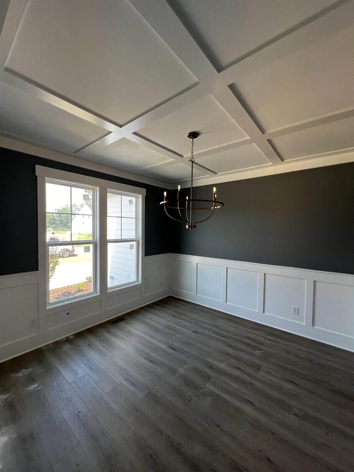 A bright dining room with dark gray walls, white wainscoting, and a coffered ceiling. A modern chandelier with six bulbs hangs from the ceiling. Large window allows natural light to fill the space. Wood flooring enhances the room's warmth.