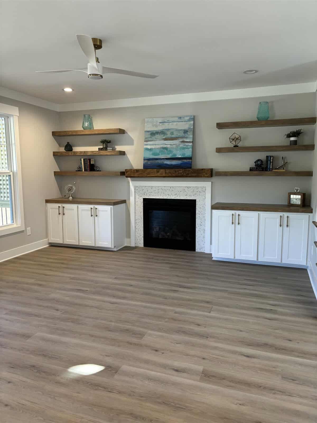 A spacious living room with light wood flooring, featuring white cabinets and wooden shelves flanking a modern fireplace. Above the fireplace hangs an abstract painting. A ceiling fan is centered above the room. Natural light enters from a window.