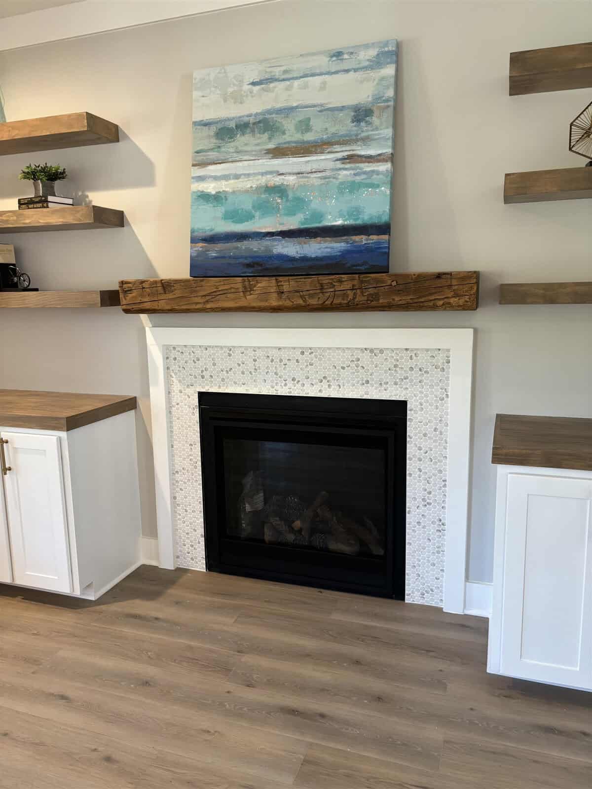 A cozy living room with a fireplace surrounded by a white mosaic tile. Above the wooden mantel sits a large abstract painting in blue and beige tones. Wooden floating shelves with decor are on either side, and the floor has light wood planks.