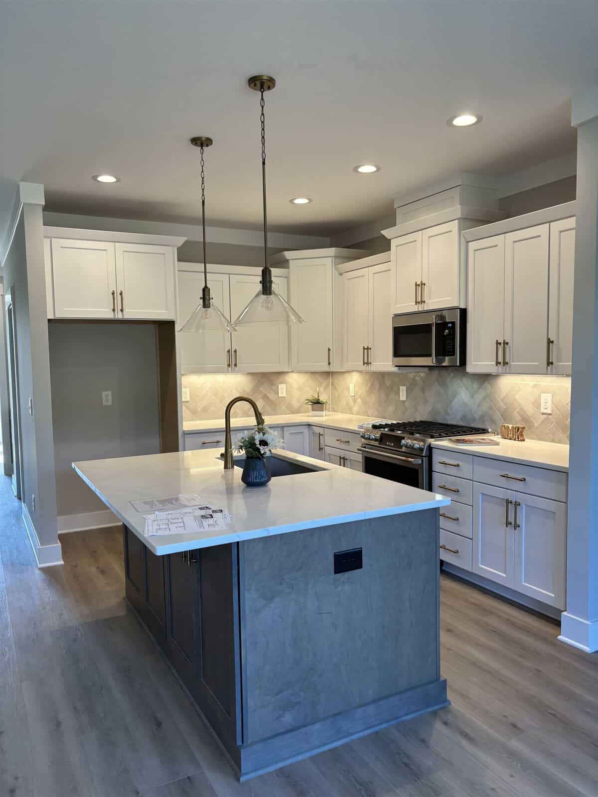 A modern kitchen with white cabinets, a gray island, wood flooring, and stainless steel appliances. Pendant lights hang over the island, and a herringbone tile backsplash accents the sleek, bright design.