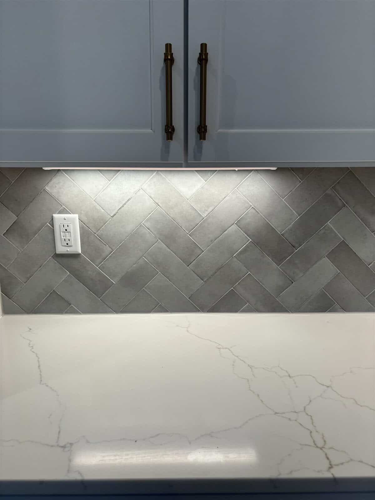 A kitchen counter features a white marble surface with subtle gray veining. The backsplash is composed of gray herringbone tiles. Above, white cabinets with gold handles are partially visible, and a power outlet is mounted on the wall.