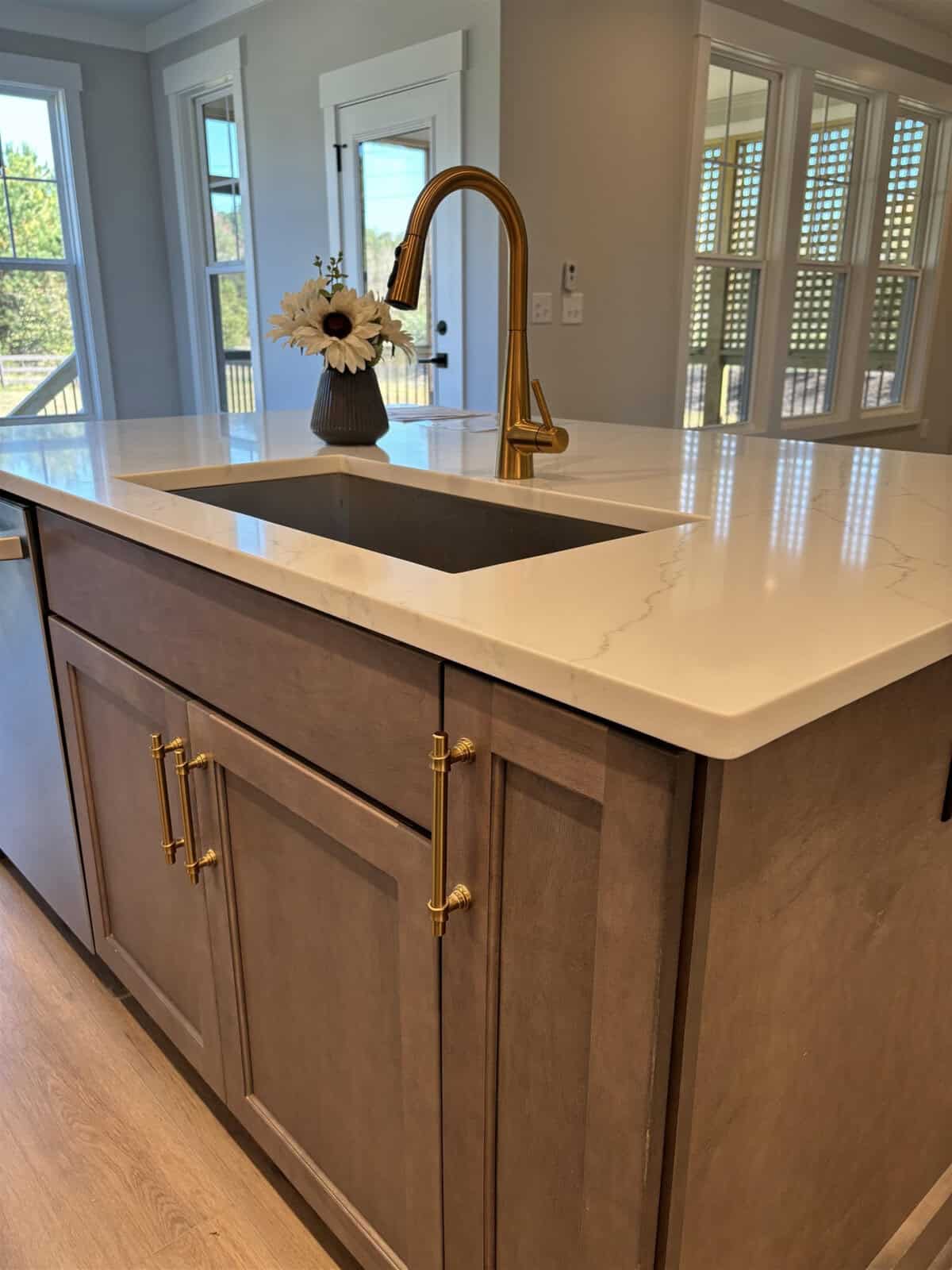 A kitchen island with a marble countertop, featuring a gold faucet over a sink. The wooden cabinetry below has golden handles. A vase with white flowers sits on the countertop. Sunlight streams through the window shutters in the background.