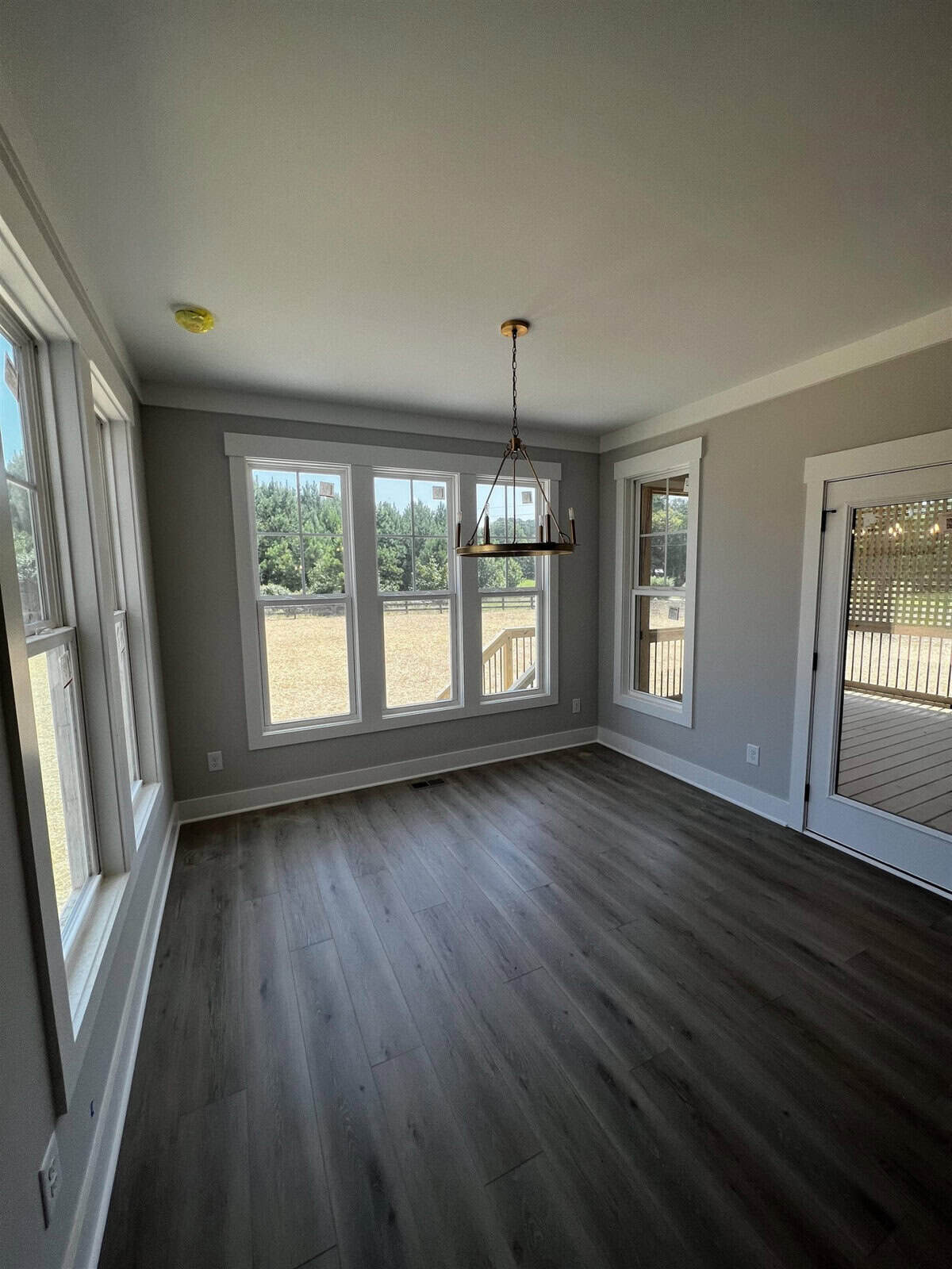 A bright, empty room with large windows allowing natural light to fill the space. The room features grey walls, dark wood flooring, a brass chandelier, and a glass door leading to a backyard or patio area.