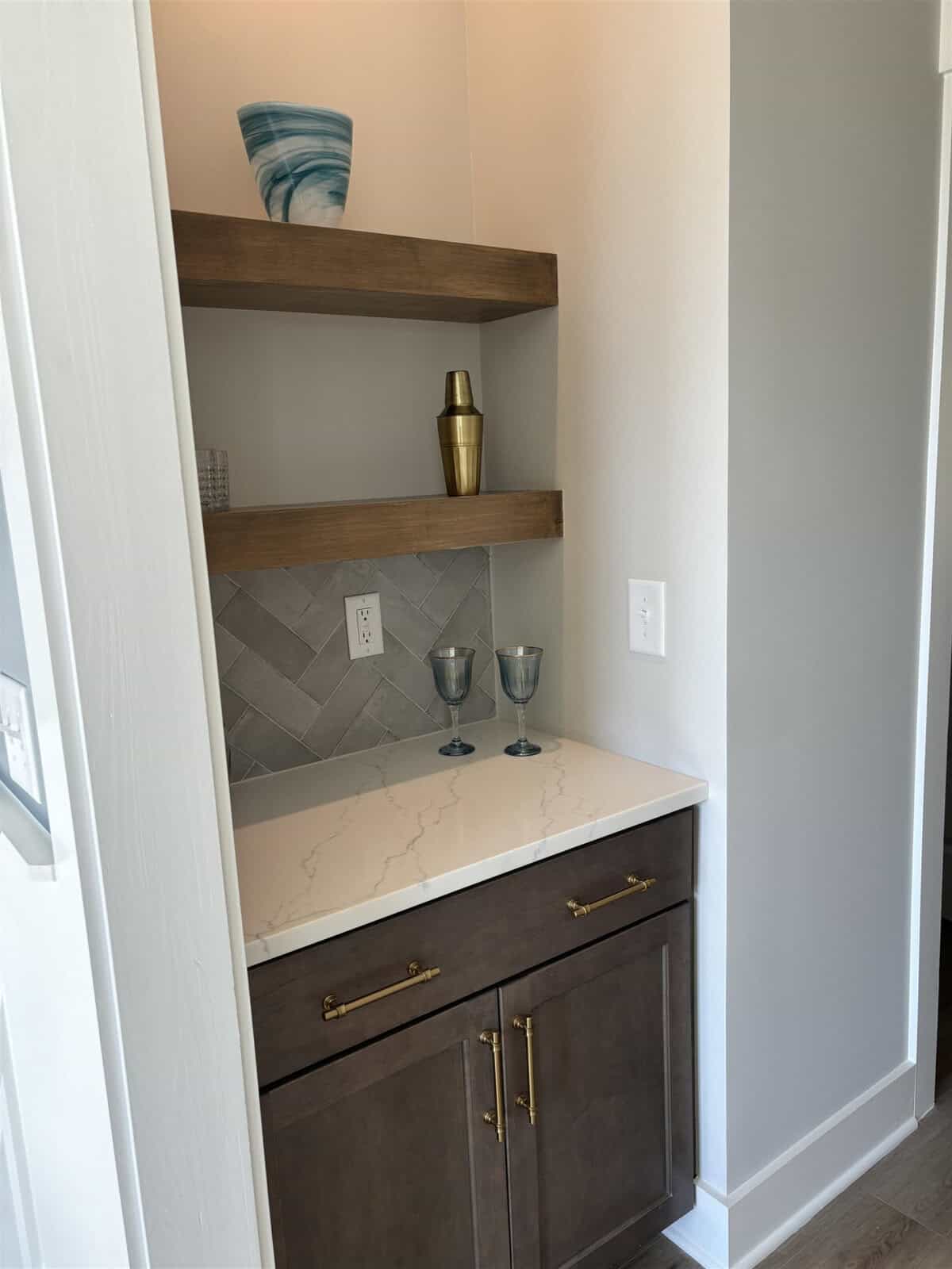 A small home bar area with a white marble countertop and a herringbone-patterned gray tile backsplash. Wooden shelves hold a decorative vase, brass shaker, and glasses. Below, a cabinet with brass handles offers additional storage.