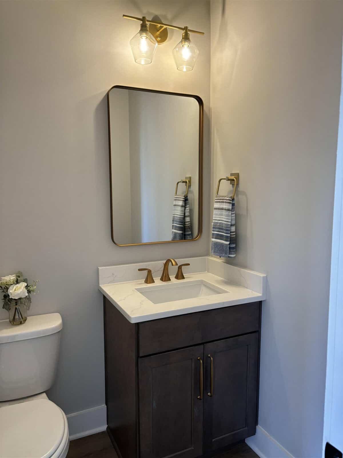 Modern bathroom with a dark wood vanity featuring a white countertop and gold fixtures. Above the sink is a rectangular mirror with rounded corners. A striped towel hangs to the right, under a dual-bulb light fixture. A small bouquet sits on the toilet tank.