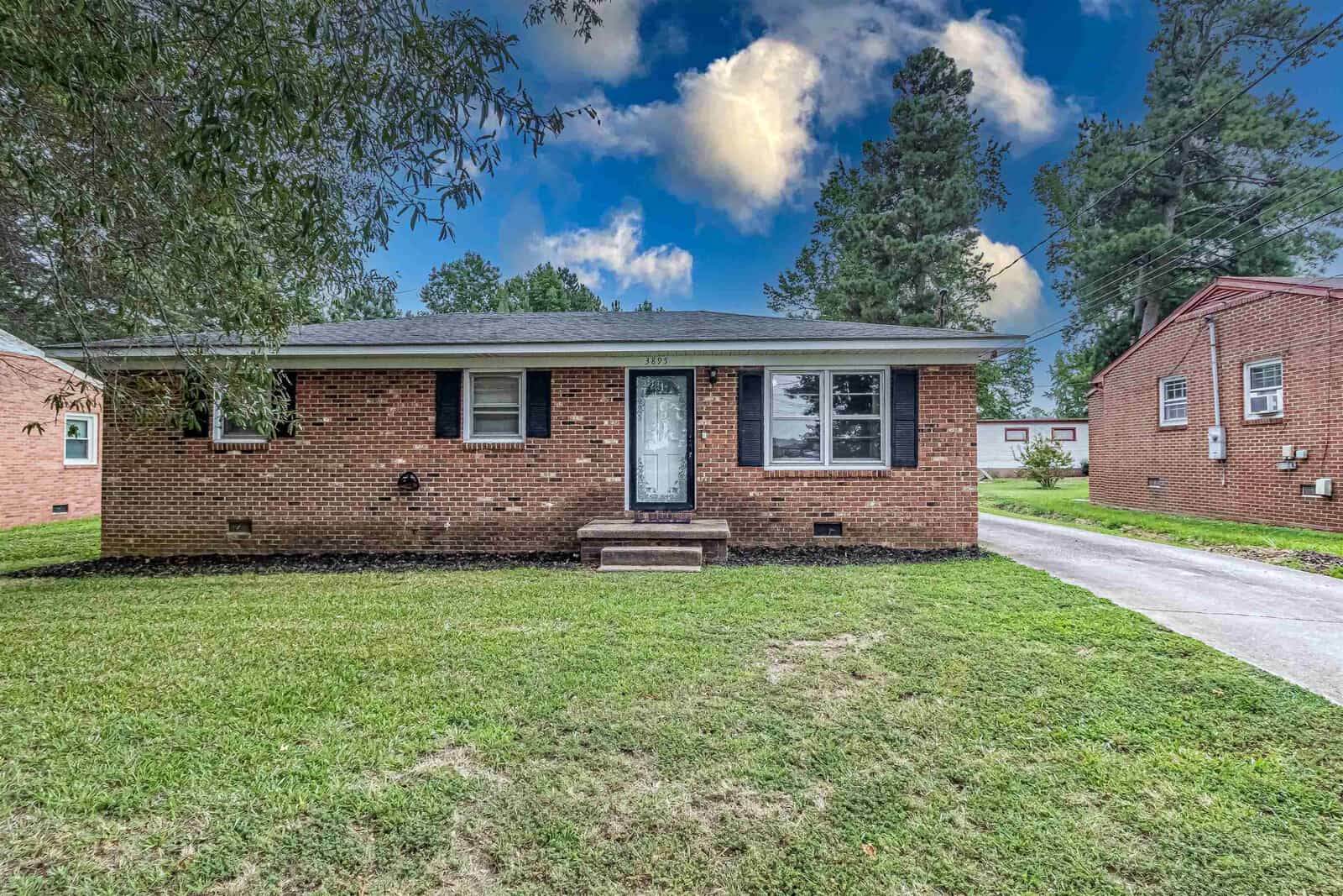 A single-story brick house at 3895 Raleigh Road, Henderson NC, with a white front door and large front window, featuring a concrete driveway on the left and a lawn in the foreground, under