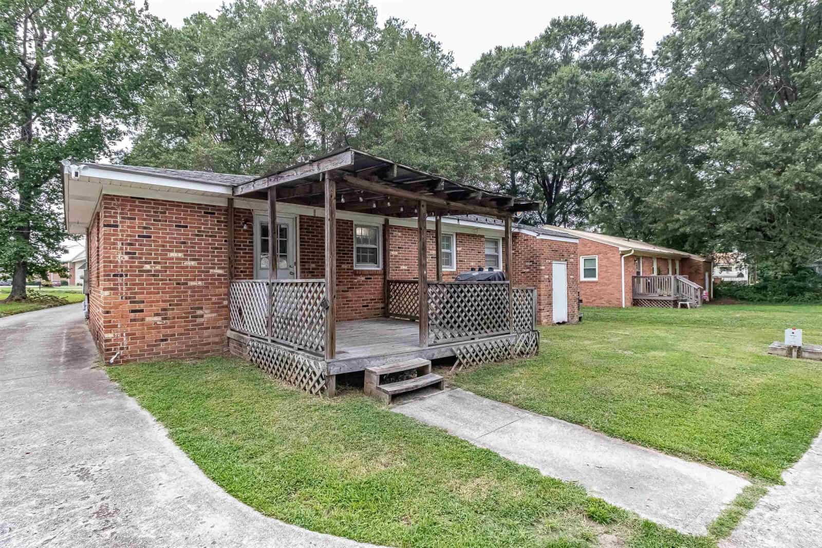 A single-story brick residential building with a wooden front porch and a latticed skirt, adjacent to a paved walkway, surrounded by grass and trees.