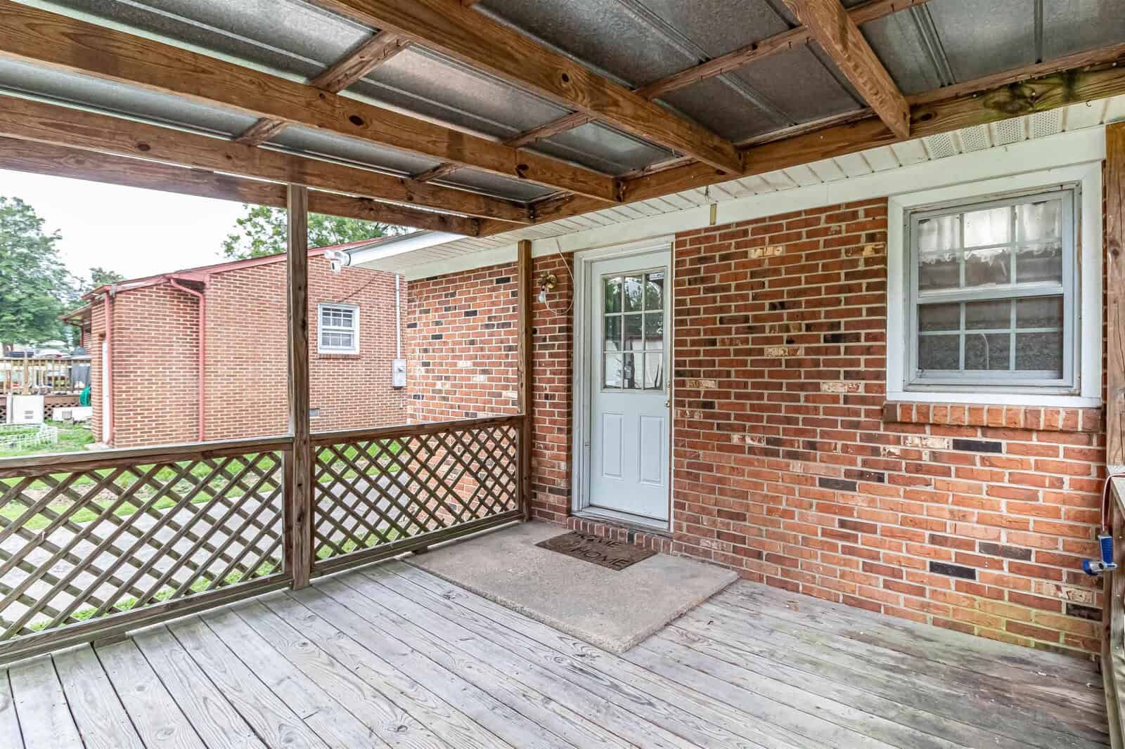 A brick house with a covered wooden porch, featuring a lattice panel, a white door, and a single window.