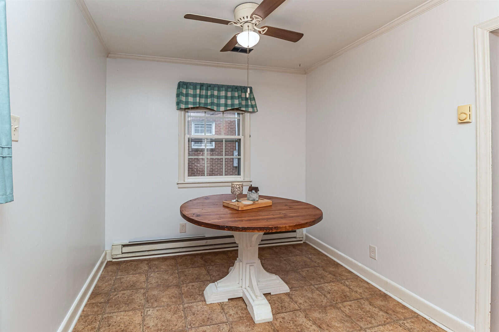 An empty room with a round wooden table under a ceiling fan, next to a window with a checkered valance curtain, and tiled flooring.