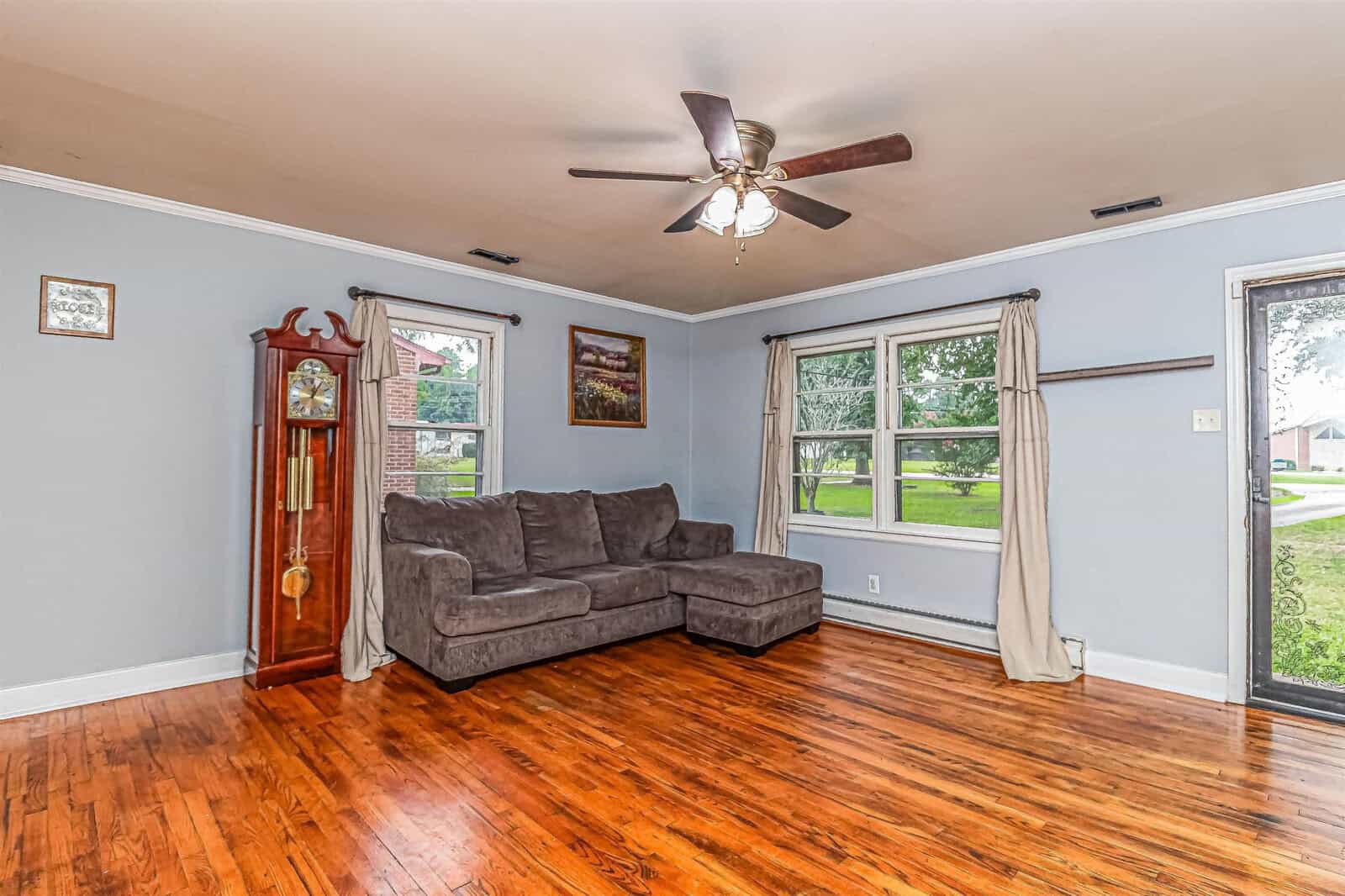 A well-lit living room with polished hardwood floors featuring a gray sectional sofa, a wooden grandfather clock to the left, and two large windows with white curtains. a ceiling fan is mounted overhead, and a few decorative items hang on the pale blue walls.
