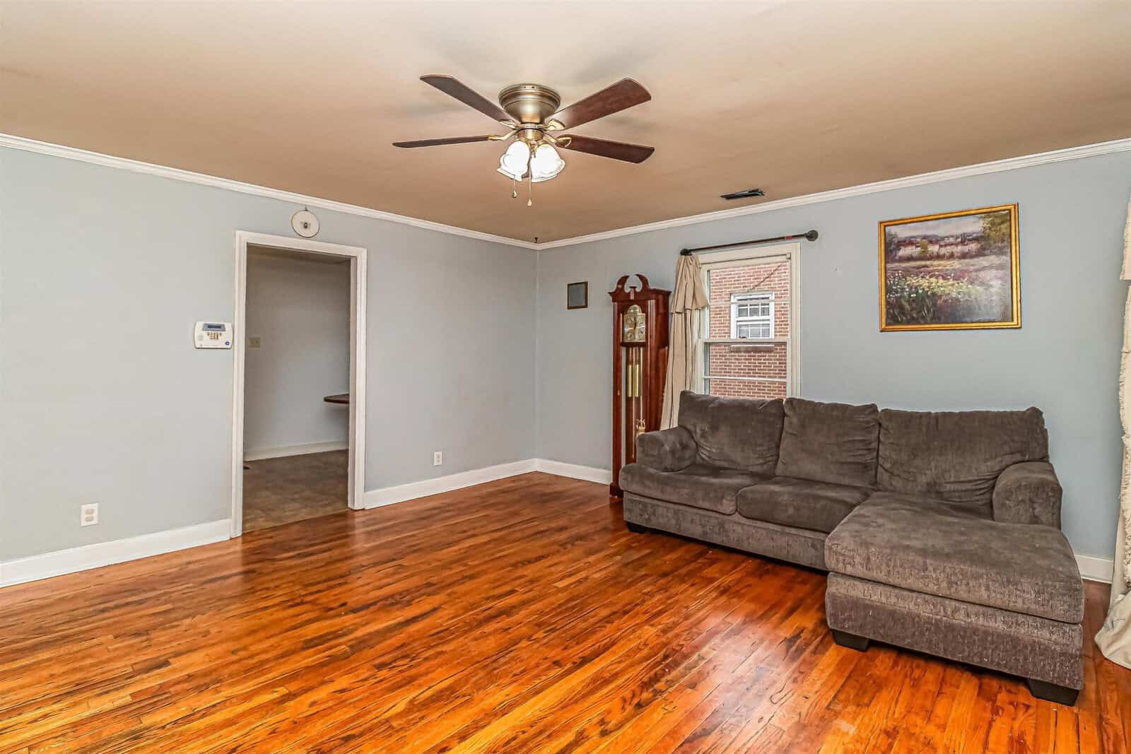 A spacious living room featuring polished hardwood floors, a gray upholstered sofa, a ceiling fan above, and a framed painting on the wall. the room has a doorway leading to another area, and there's a grandfather clock standing to the right.