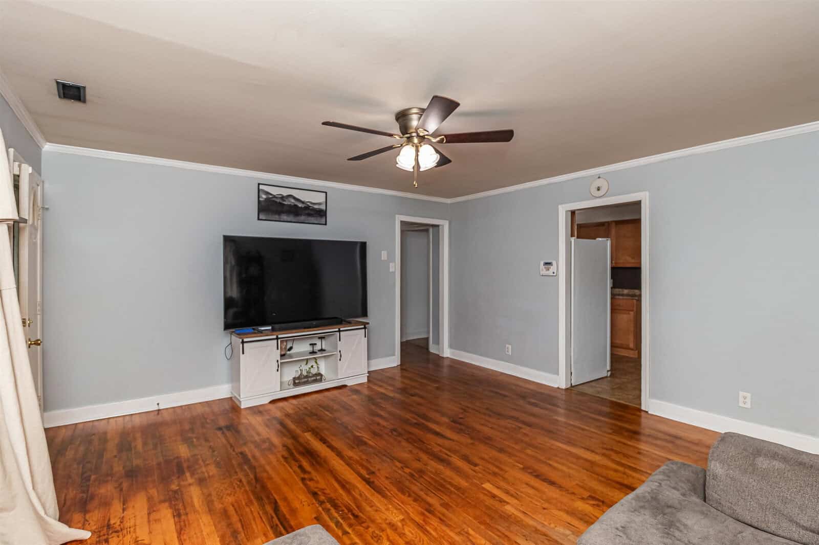 A spacious living room with glossy hardwood floors featuring a ceiling fan, a flat-screen tv on a glass stand, light blue walls, and an open doorway.