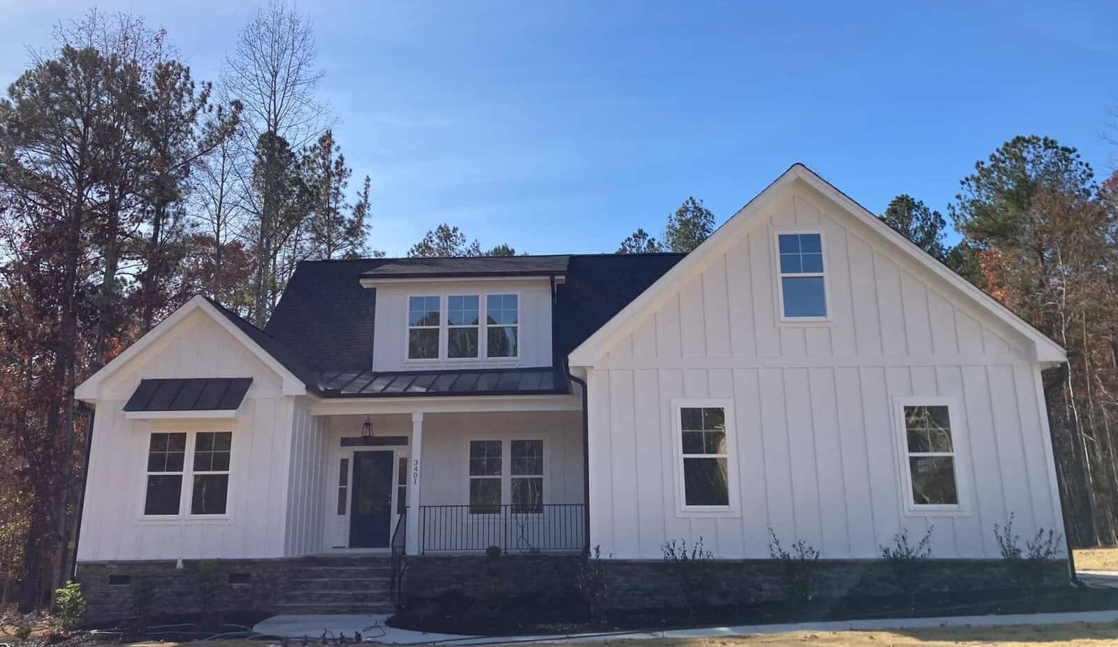 Situated at 3401 River Manor Court, this white modern farmhouse-style home in Franklinton boasts a sleek black roof. A large front porch and numerous windows open up to a wooded area with tall trees, while the clear blue sky provides a stunning backdrop.