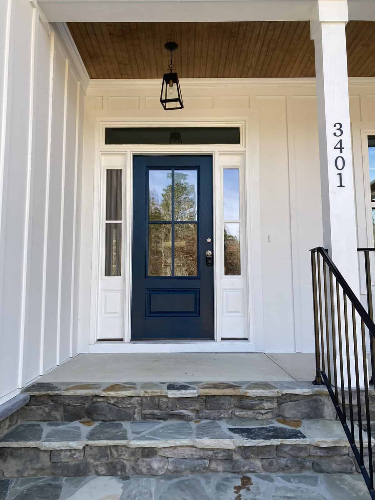 A dark blue front door with glass panels is set in a white house, flanked by narrow windows. Stone steps lead up to the door, and a black metal railing runs alongside. A black pendant light hangs above, and the house number "3401" is displayed vertically.