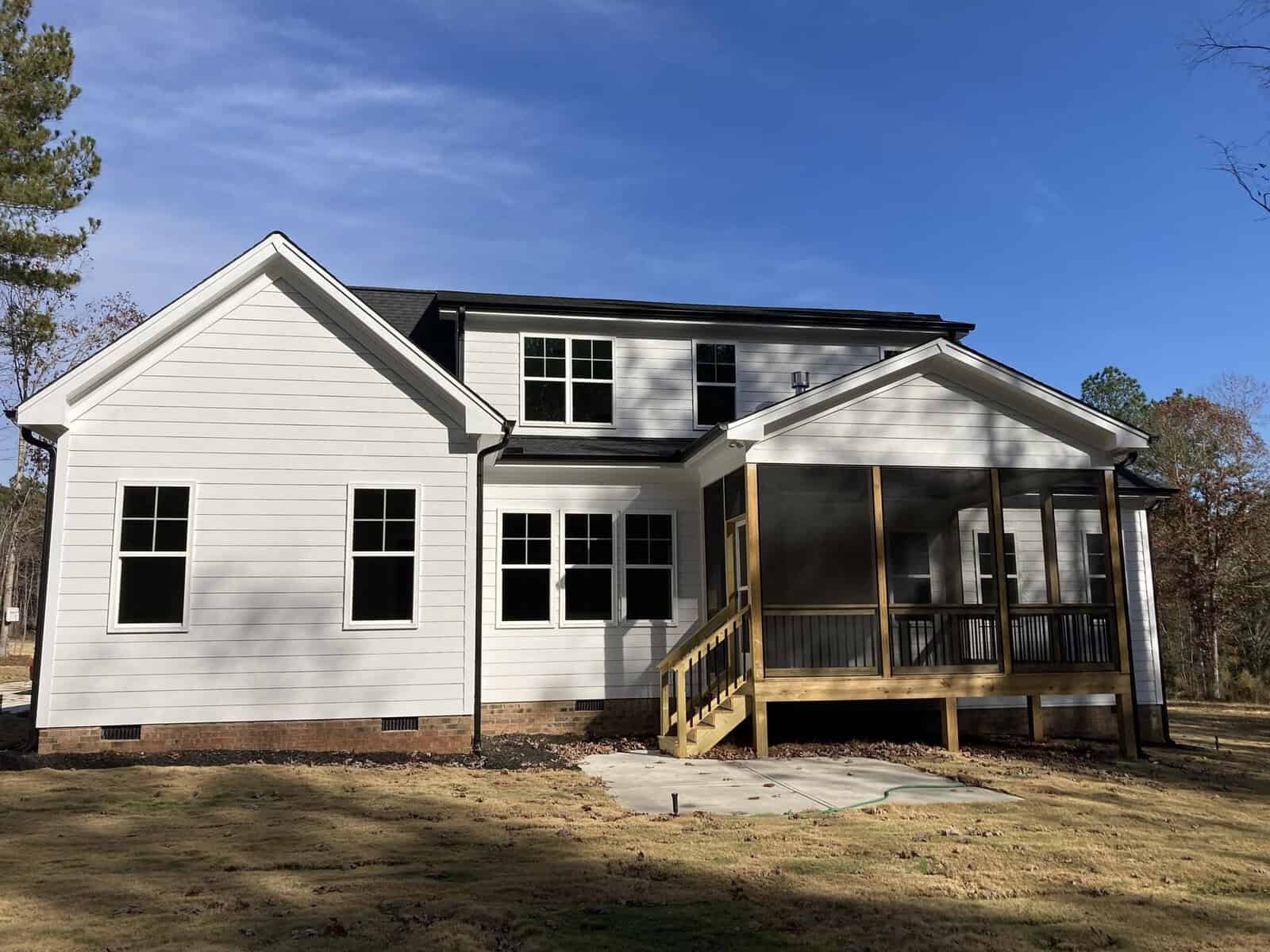 A two-story white house with black trim and multiple windows. It features a wooden screened-in porch with stairs leading to a grassy backyard under a clear blue sky. Trees surround the area.
