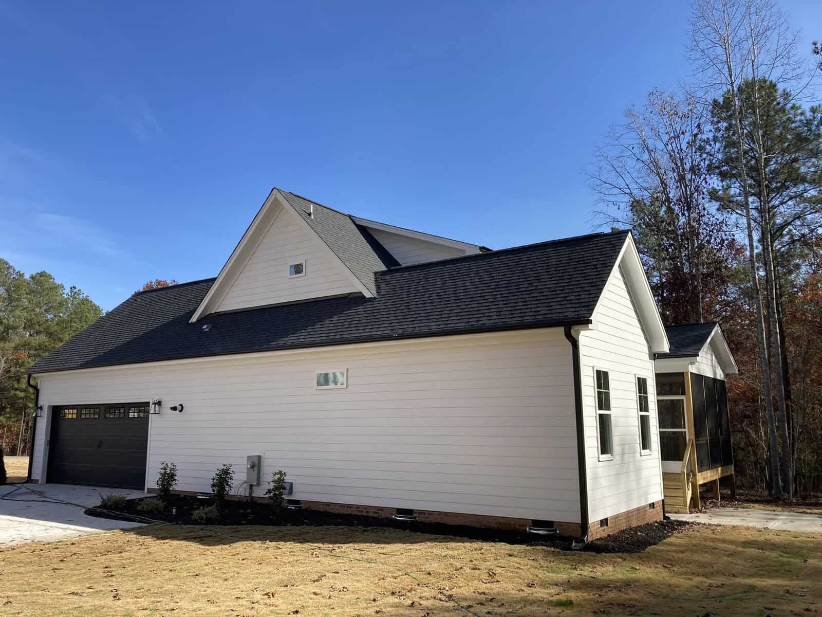 A modern, single-story white house with a dark roof, seen from the side. It has a driveway leading to a garage with a dark door. The yard is grassy, and there are sparse trees in the background under a clear blue sky.