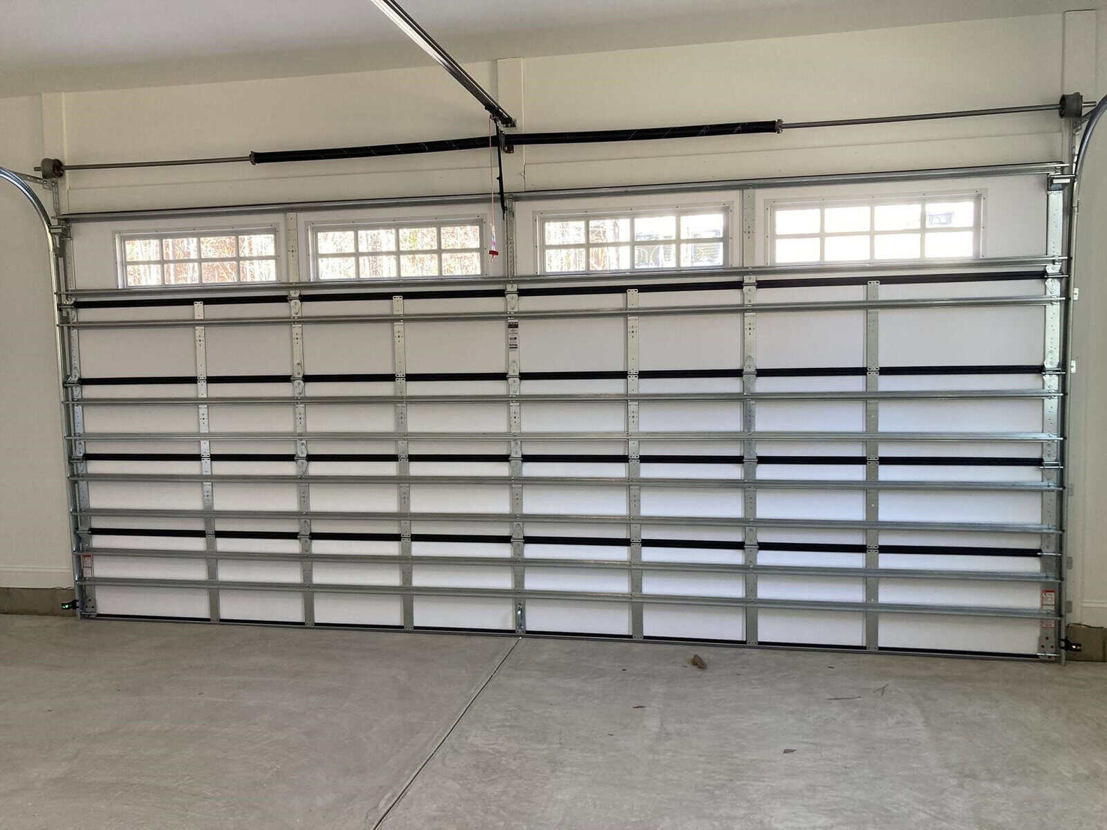 A closed, double-wide garage door with small, rectangular windows at the top. The floor is concrete, and the space appears to be well-lit, reflecting natural light from the windows.