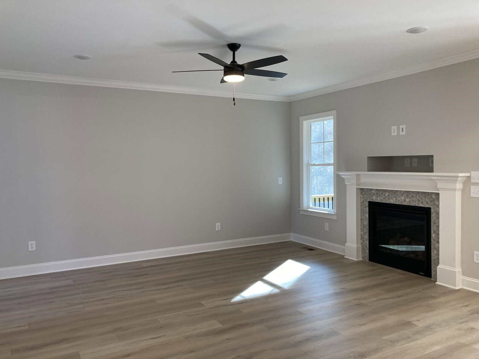 A spacious, empty living room with light gray walls and wood flooring. A modern ceiling fan is centered above. A fireplace with a white mantel and gray tiling is on the right, next to a window letting in natural light.