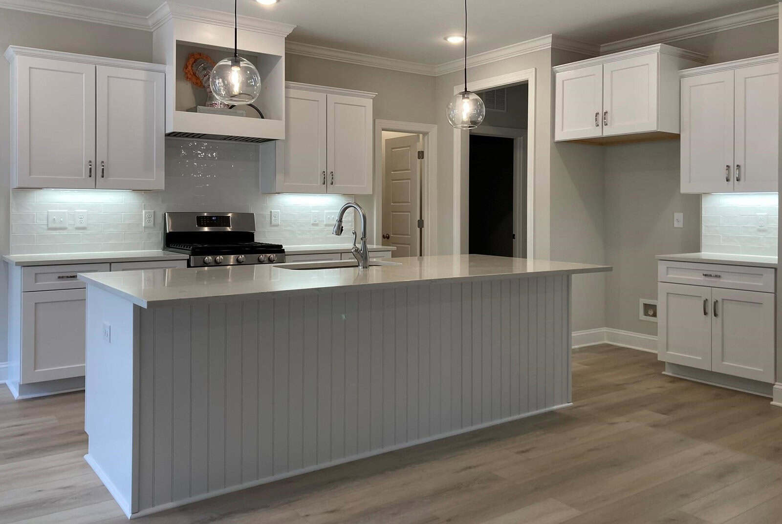 A modern kitchen with white cabinets, a large island with a ribbed design, and pendant lights. Stainless steel appliances include a stove and oven. Light wood flooring complements the contemporary design.