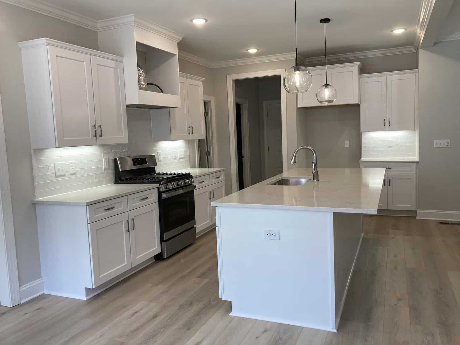 Modern kitchen with white cabinets, stainless steel appliances, and a large central island with a sink. Pendant lights hang above the island. Light wood flooring and recessed lighting create a bright, clean atmosphere.