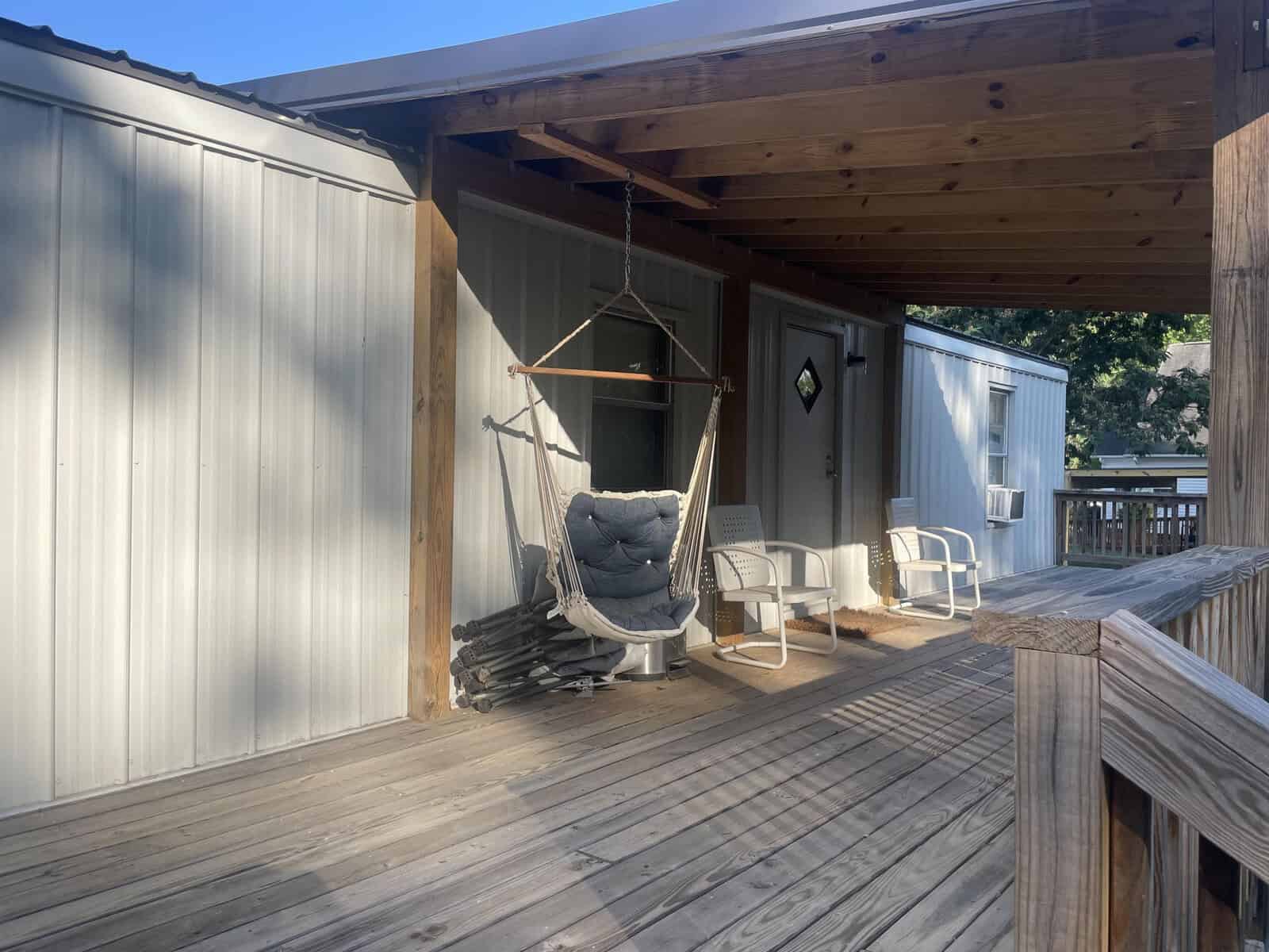 A wooden porch with a hanging chair and two additional chairs on a sunny day, with the entrance to a building in the background.