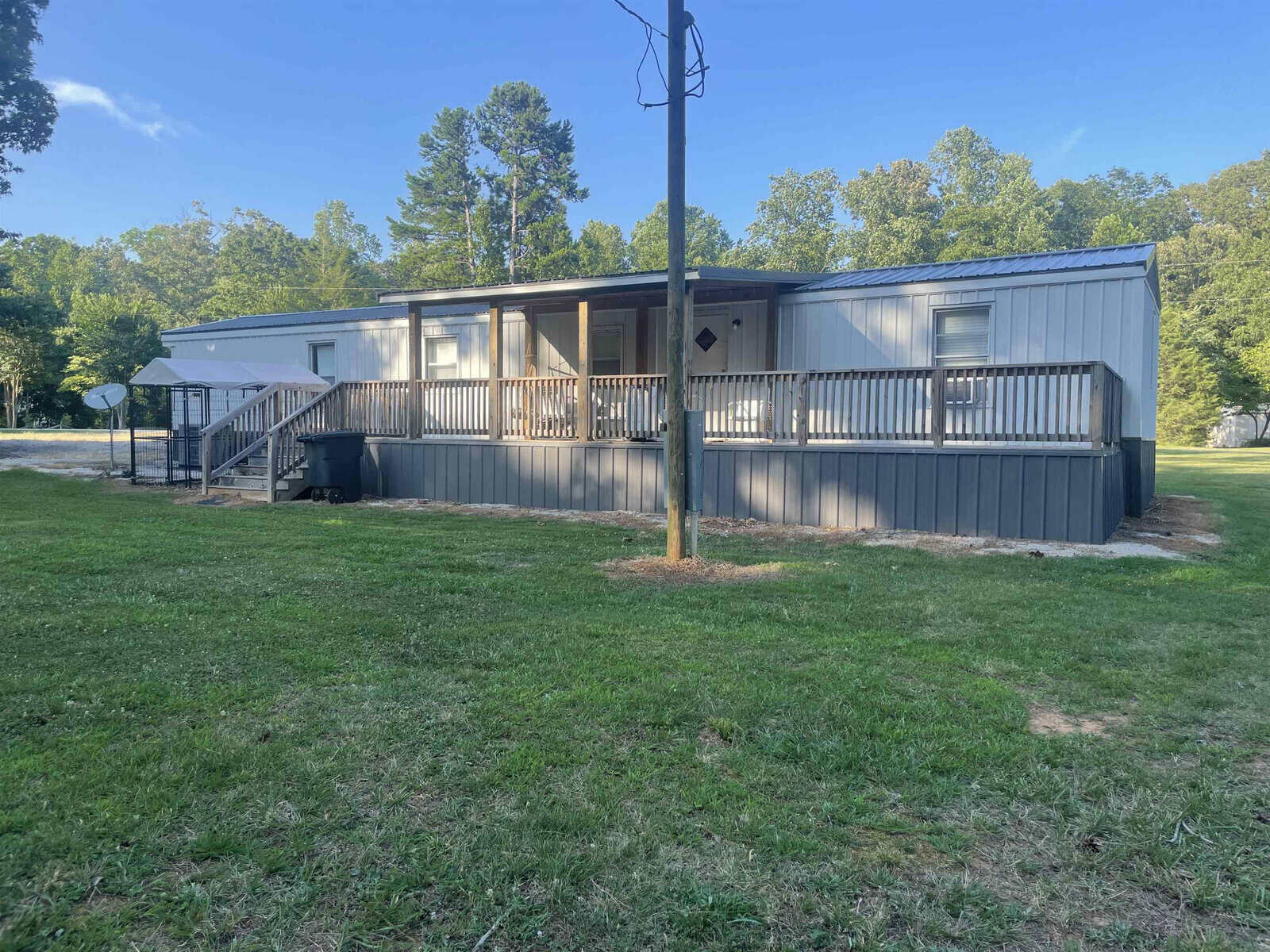 A single-story gray mobile home with a front porch and wooden steps, surrounded by a grassy lawn with trees in the background, under a clear blue sky.