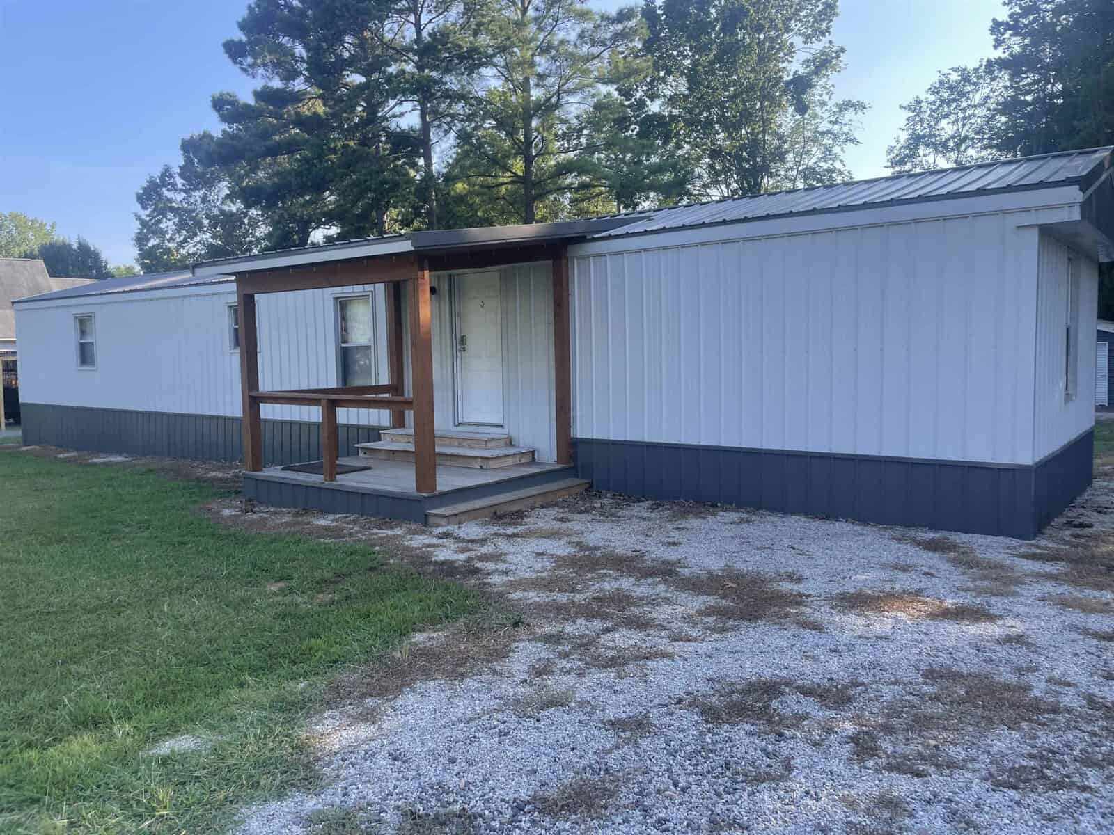 A single-story mobile home with gray siding and white trim, featuring a small wooden porch with steps and surrounded by a gravel-covered ground with a backdrop of trees and a clear sky.
