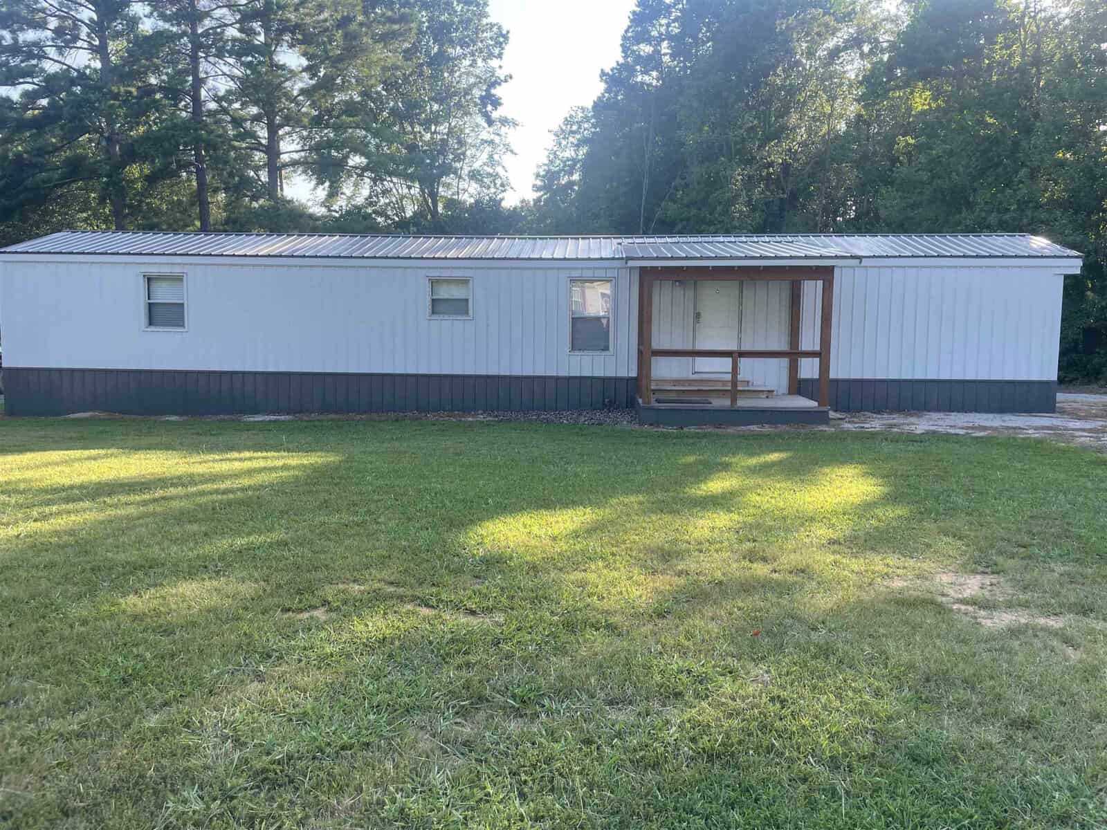 A single-story gray mobile home with a wooden front porch situated in a grassy area with trees in the background during daylight.