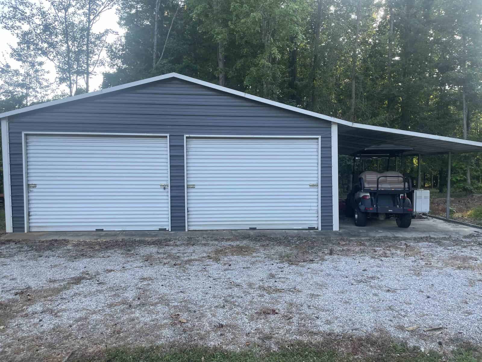 A metal double garage with closed doors and an attached carport, under which is parked a golf cart, surrounded by trees.