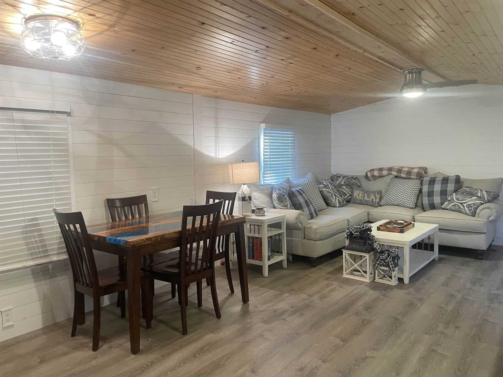 A cozy, well-lit interior of a living space featuring a wooden dining table with chairs, a comfortable sofa with cushions, and a white wood-paneled wall with a sloped ceiling.
