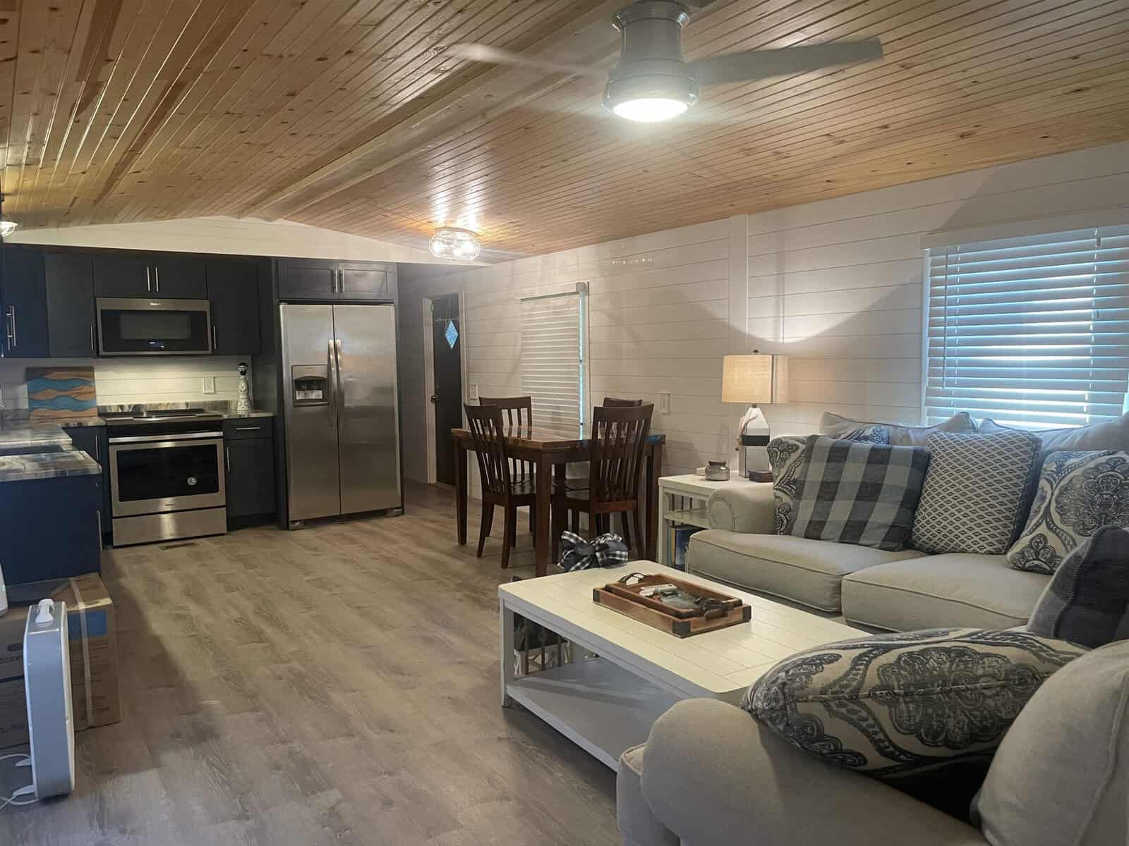 Interior of a modern living space featuring a kitchen with stainless steel appliances, a wooden dining table set, and a living area with a plush sofa and patterned cushions, under a wooden ceiling with a fan.