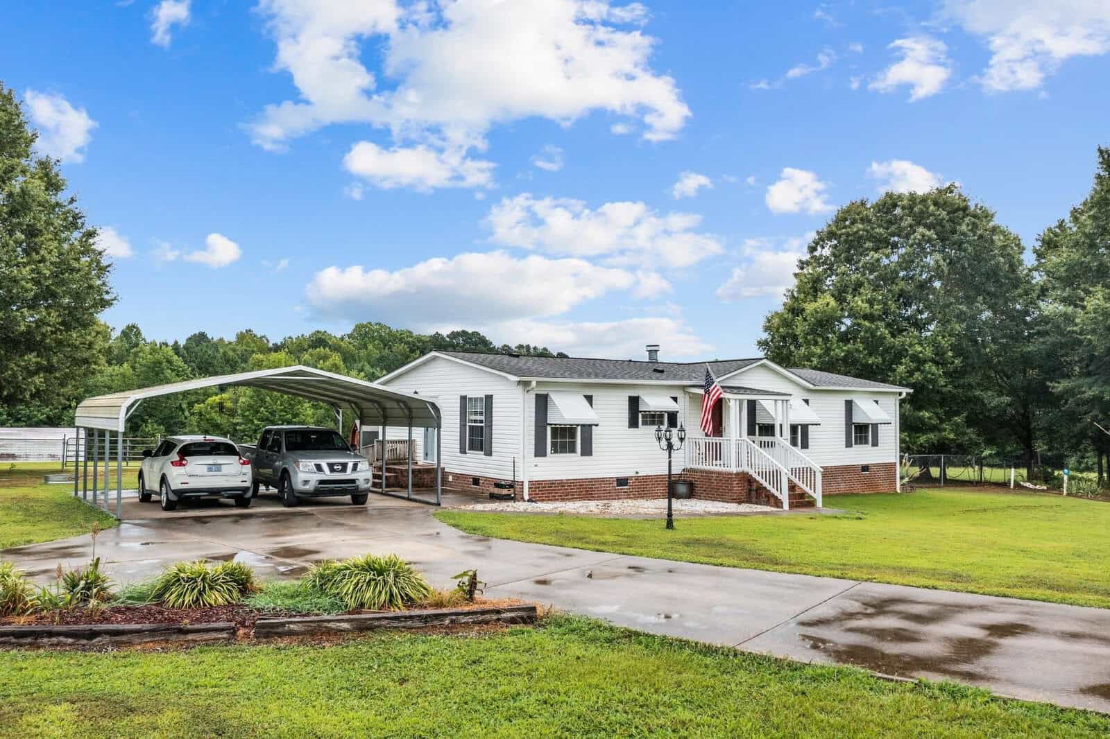A single-story white house with a metal carport sheltering two vehicles, located at 151 Southerland Mill Road, Henderson NC, is set against a backdrop of green lawn and trees under a