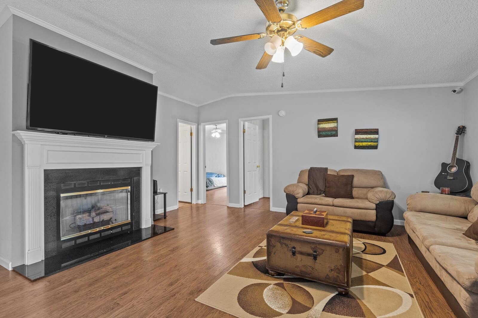 A well-lit living room featuring a fireplace, a ceiling fan, and a flat-screen tv, furnished with a brown sofa, a loveseat, a vintage trunk coffee table, a guitar in the corner, and decorative items on the wall.