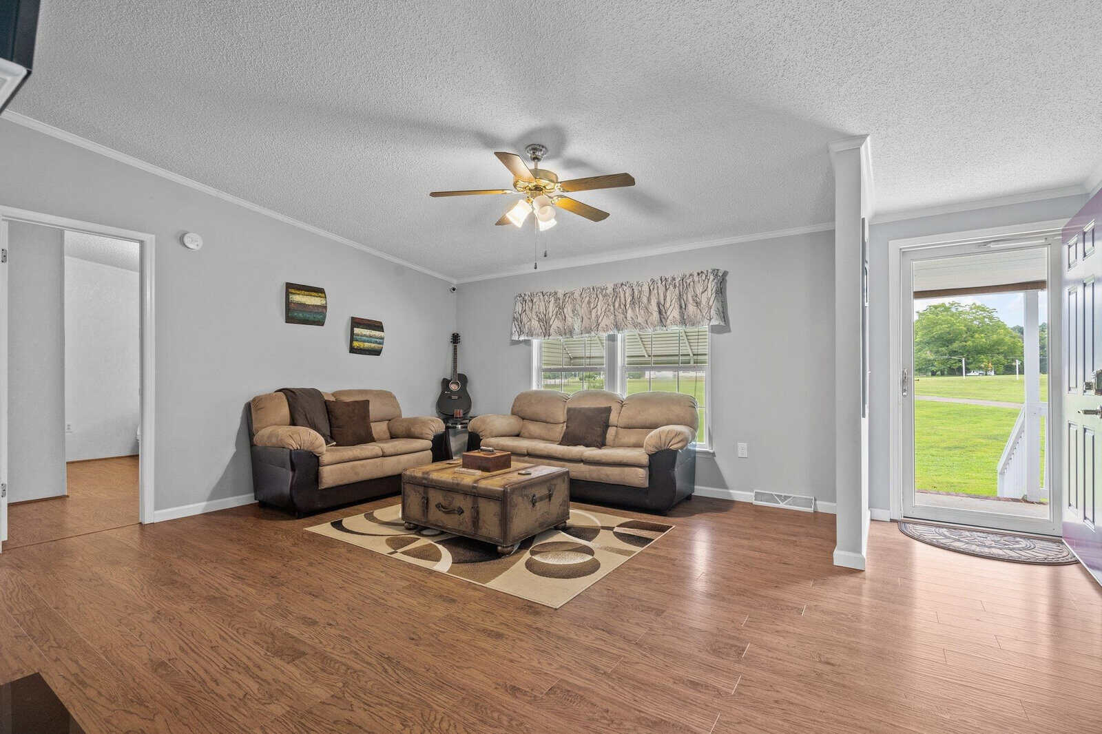 A well-lit, tidy living room with a two-seater sofa, a matching armchair, and an ottoman set on a beige rug. the room features laminate flooring, white walls with minimal artwork, a ceiling fan with lights, and a large window with a rolled blind partially revealing an outdoor view. an open doorway leads to a brightly illuminated adjacent area.