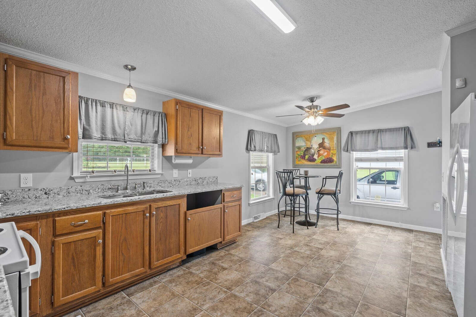 A well-lit kitchen interior with wooden cabinets, granite countertops, a ceiling fan, tiled flooring, and a breakfast bar with stools near a window.