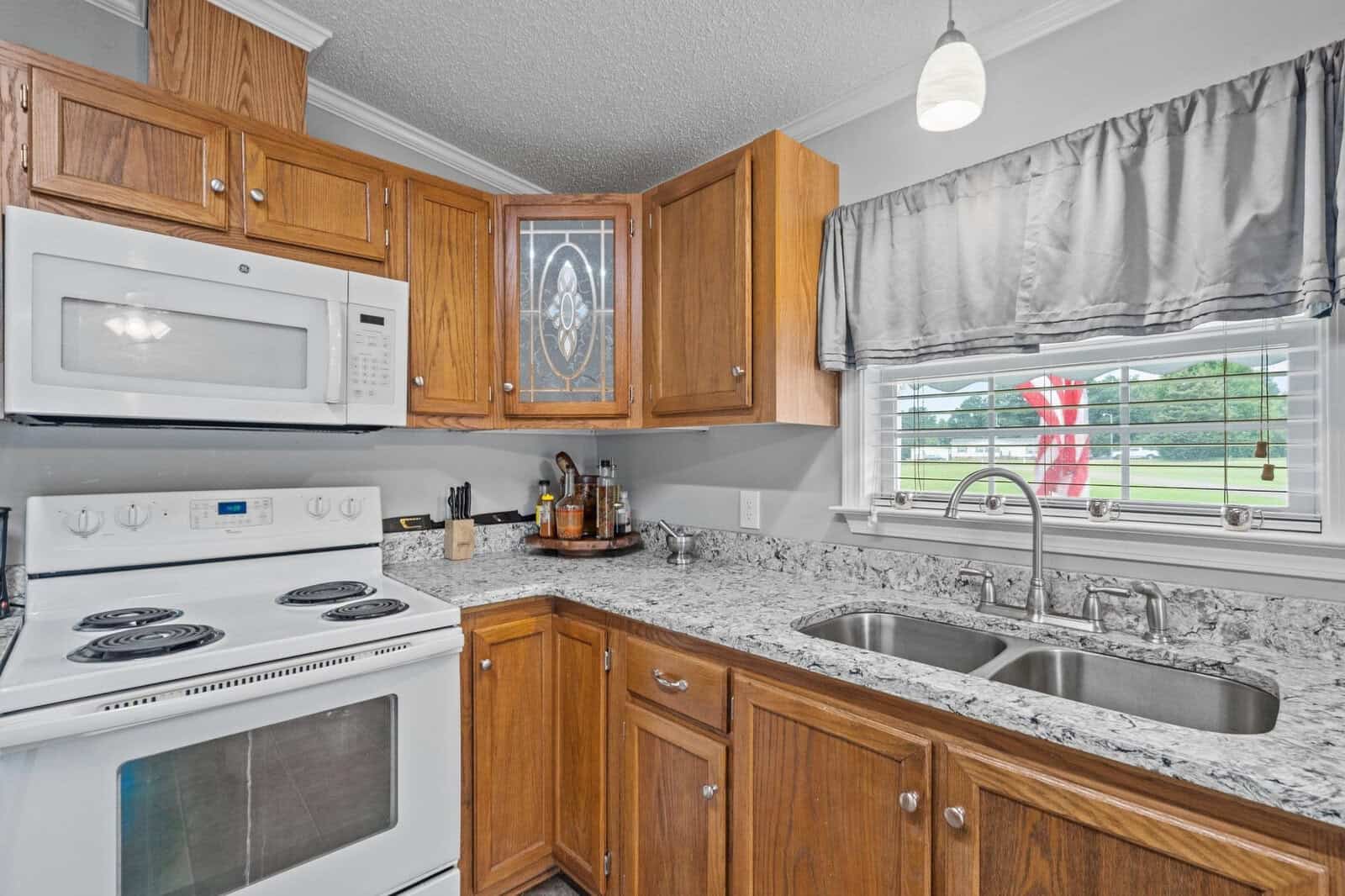 A modern kitchen featuring stainless steel appliances, granite countertops, and wooden cabinetry with a window dressed in gray patterned curtains looking out to a green yard.