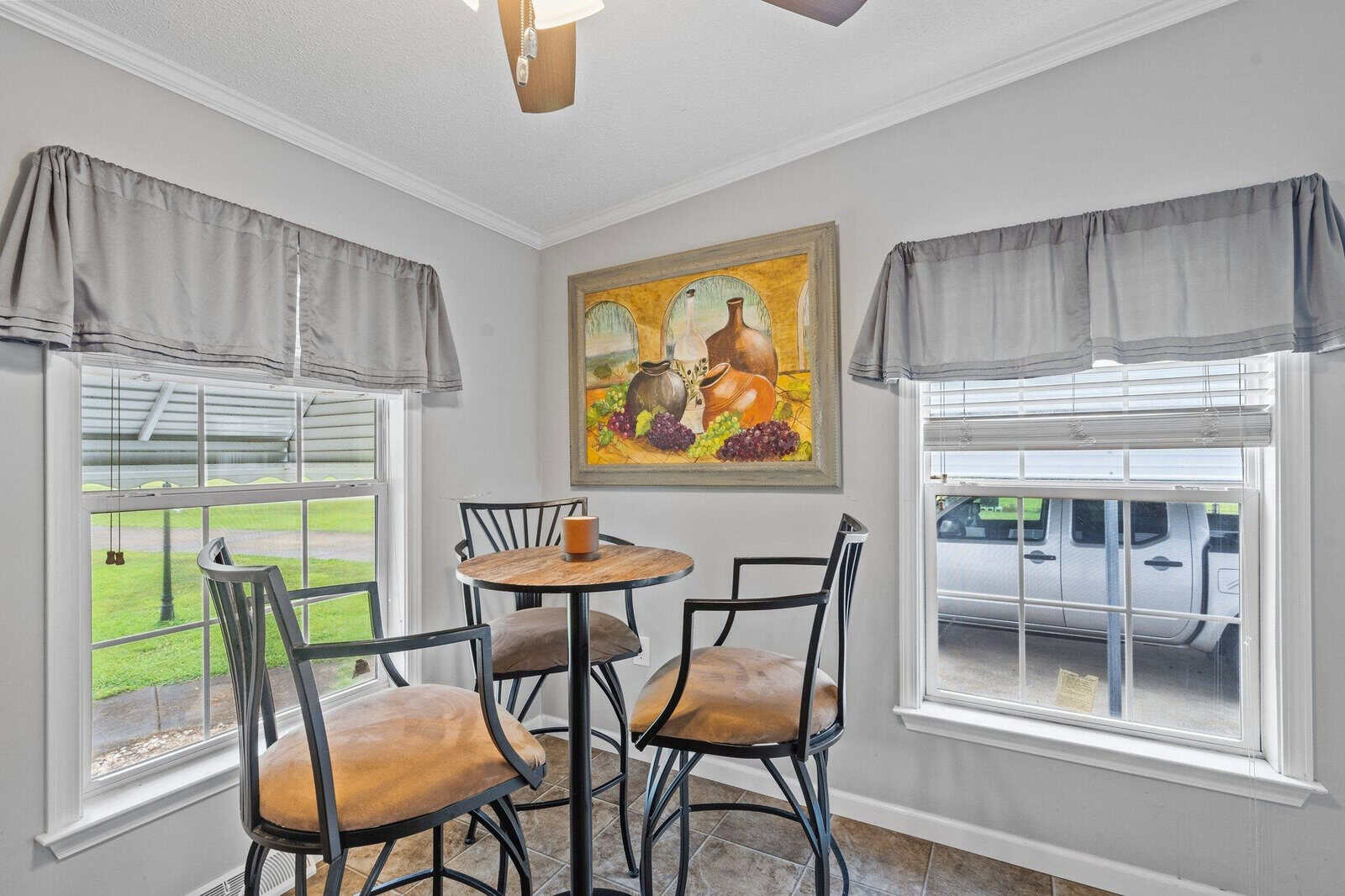 A small, neatly arranged breakfast nook with a round table and four high chairs, flanked by two windows with gray valances, and a framed still-life painting hanging on the wall.