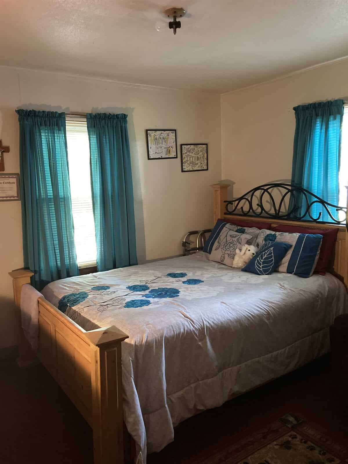 A neatly made bed with a blue and white theme in a well-lit bedroom with matching curtains and decorative pillows, complemented by wall art and a small area rug.